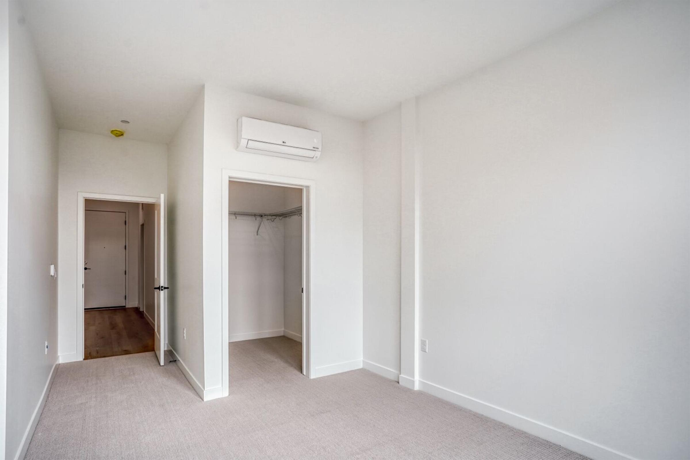 Empty room with light-colored walls and carpeted flooring. One door leads to a closet with shelving, and another door is partially visible, suggesting a hallway. A wall-mounted air conditioning unit is installed above the doorway. Natural light filters into the space, creating a bright ambiance.