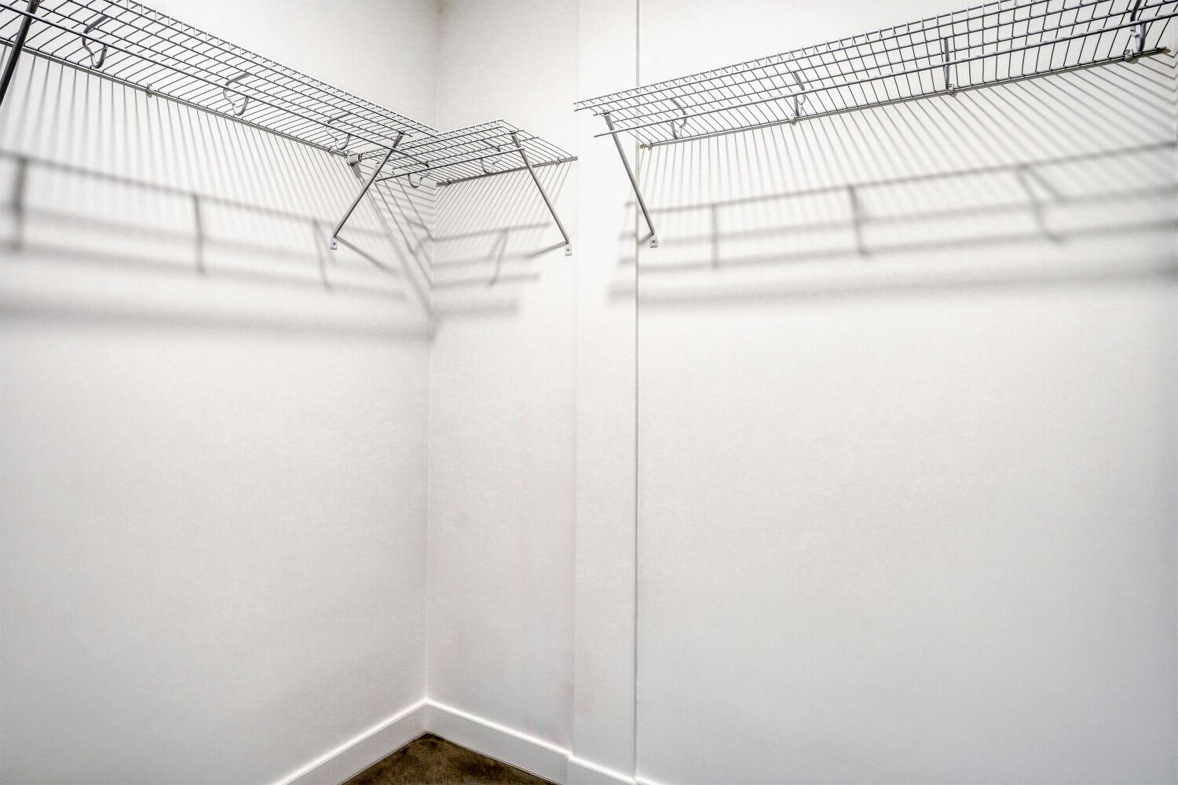 Empty closet space with two wire shelves mounted on the wall. The shelves are casting shadows on the plain white walls, and there is a blank floor with light-colored tiles. A mirror is visible on one wall, reflecting the shelves.