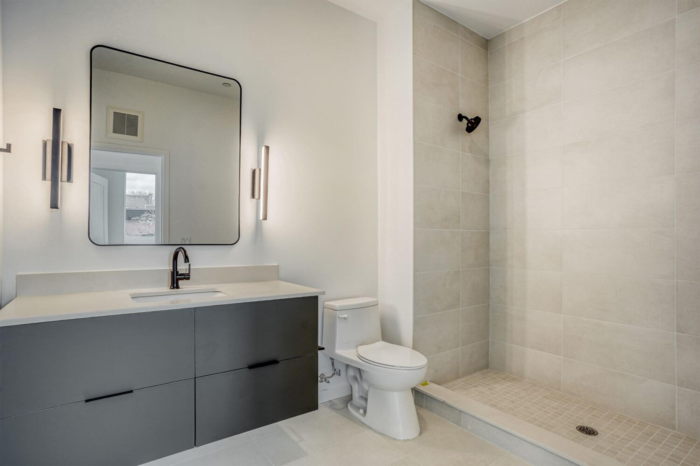 Modern bathroom featuring a sleek design. It includes a large mirror, a simple sink with a dark cabinet underneath, a toilet, and a walk-in shower area with beige tiles. Soft lighting adds a warm ambiance to the space.
