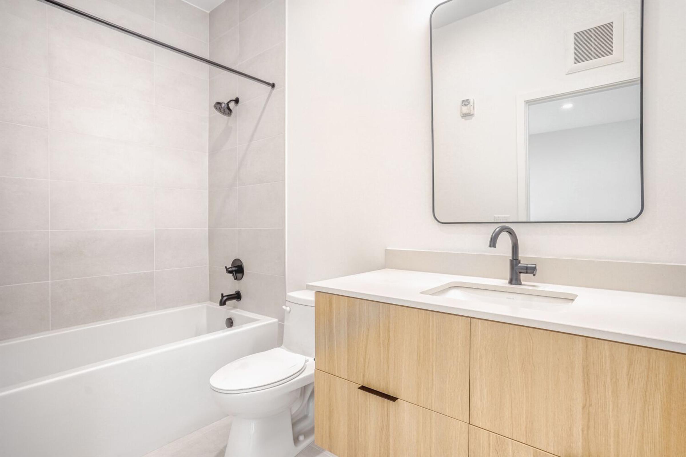 A modern bathroom featuring a light gray tiled wall, a bathtub with a showerhead, a sleek sink with a minimalist faucet, and a wooden vanity with a black handle. A round mirror is mounted above the sink, and there is a toilet nearby. The space has a bright and clean aesthetic.
