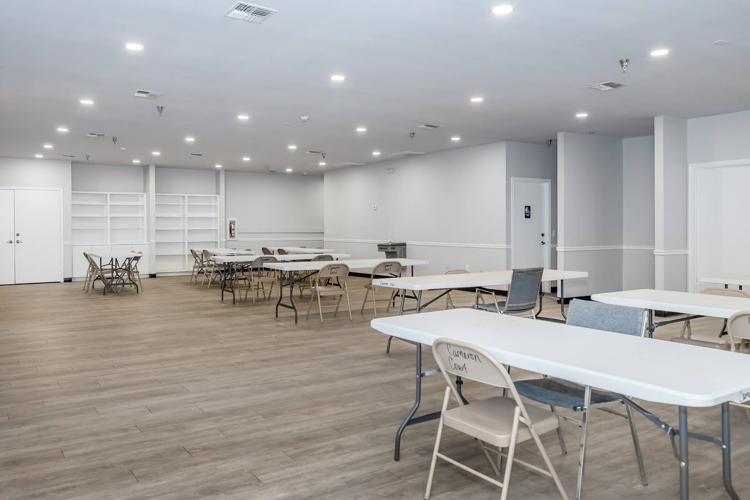 A spacious, empty meeting room featuring several tables and chairs arranged neatly. The floor is wooden, and the walls are painted light gray. There is a shelf along one wall and a doorway leading to another area. The lighting is bright, creating an open and inviting atmosphere.