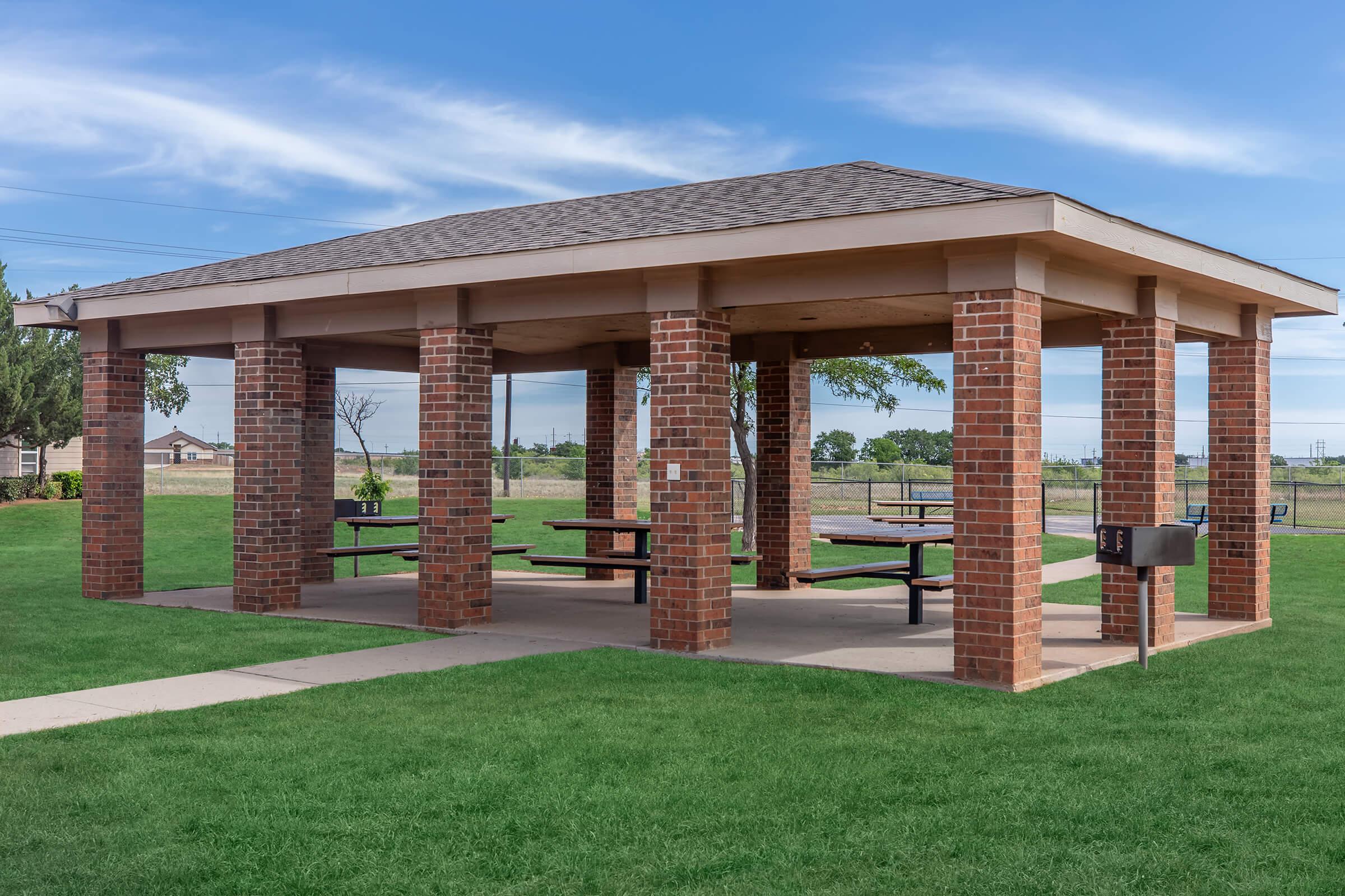 A brick pavilion with a sloped roof, featuring several picnic tables underneath, set in a grassy area. The structure has open sides, allowing for a view of the surrounding landscape, which includes trees and a clear blue sky.