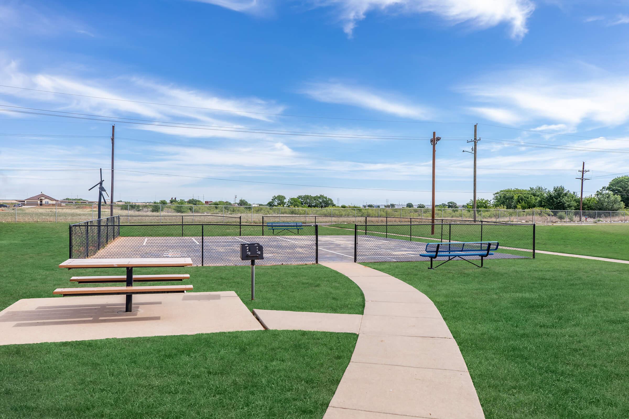 A view of a park featuring a paved path leading to a fenced tennis or basketball court, with a picnic table in the foreground. The background shows open grassland under a blue sky with clouds, and utility poles in the distance.