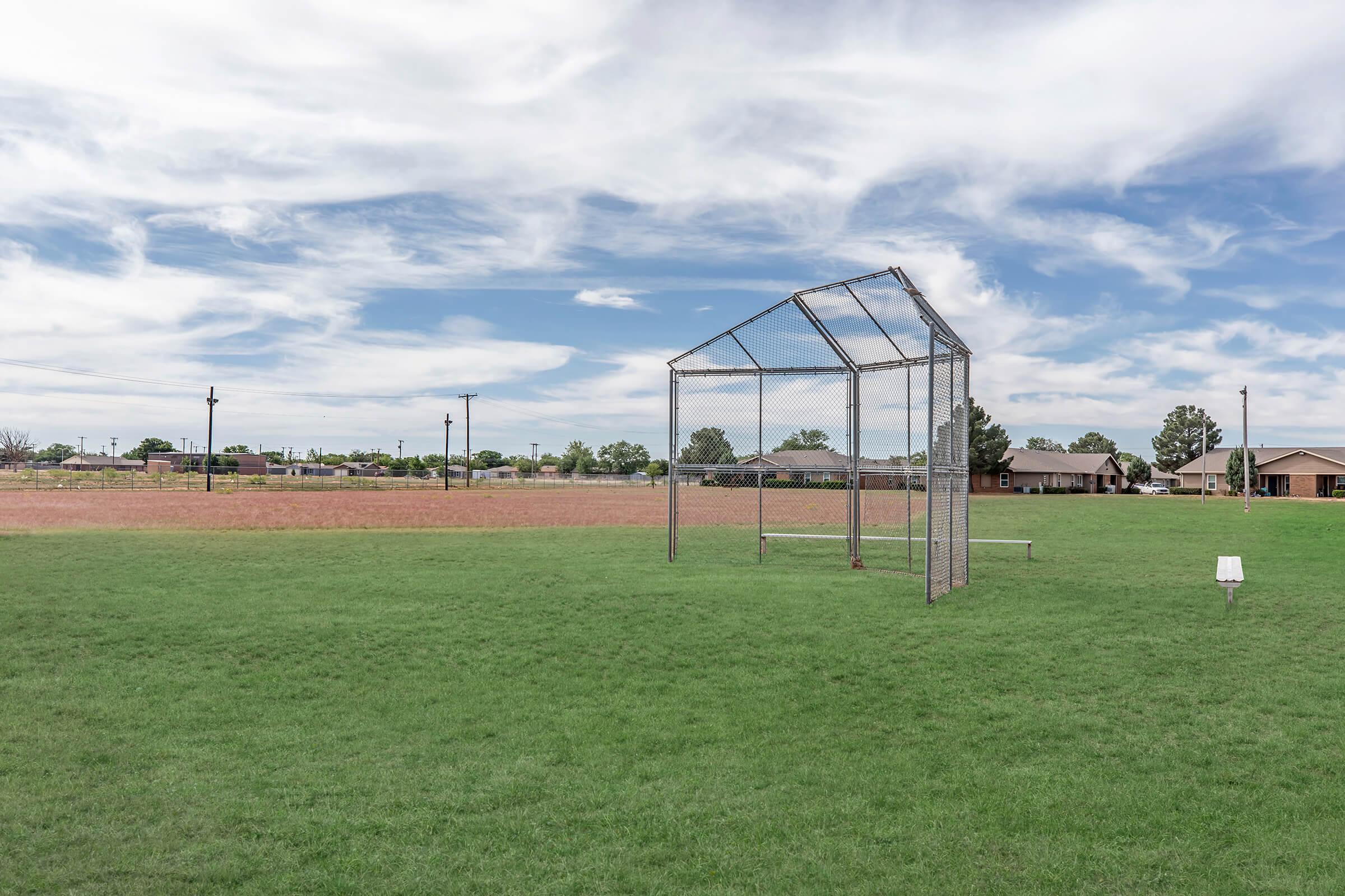 An empty baseball batting cage on a grassy field, surrounded by open space and houses in the background. The sky is partly cloudy, with blue and white clouds creating a serene atmosphere. A dirt area is visible beyond the grass, suggesting a multi-use outdoor space.