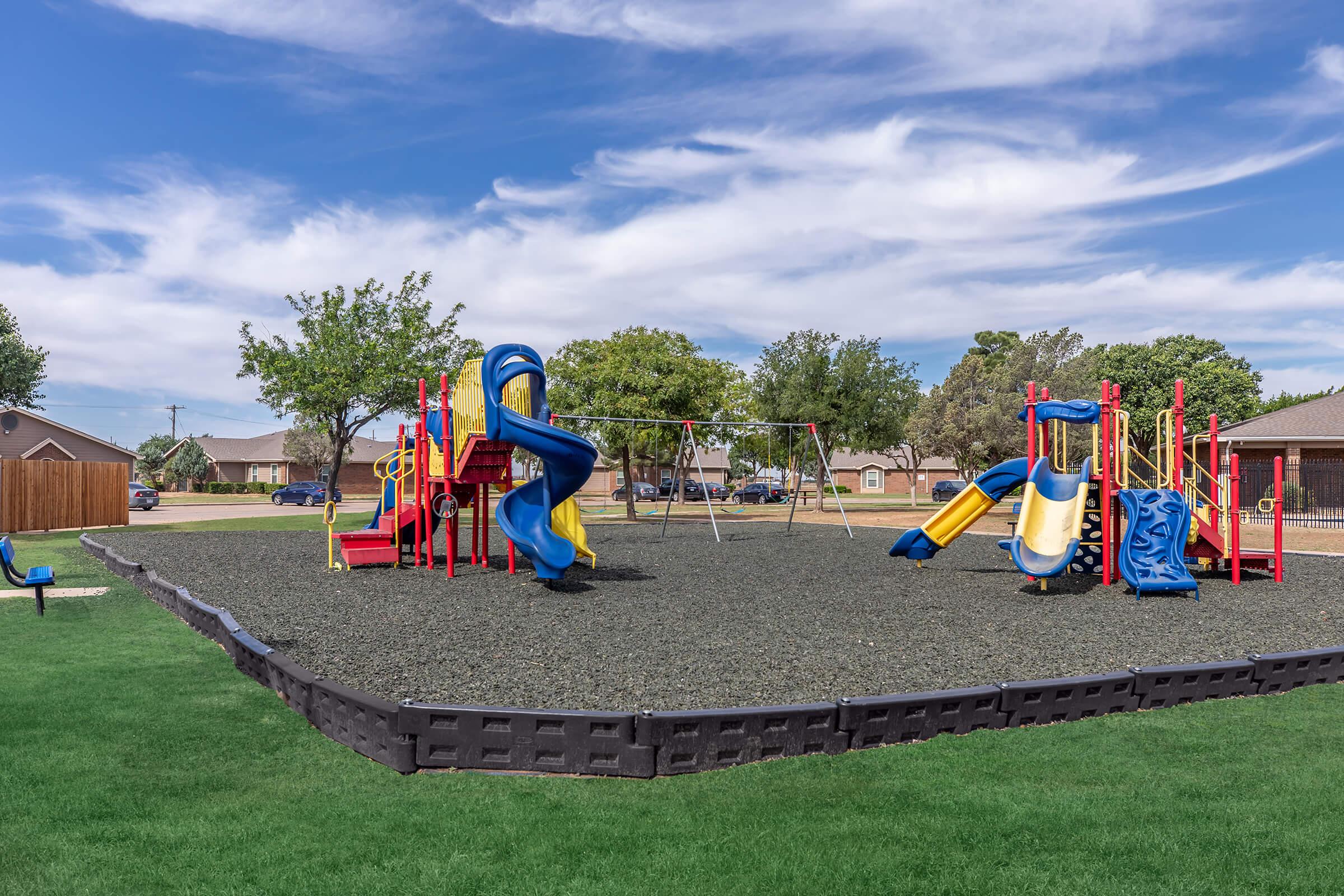 A bright, colorful playground featuring multiple slides, swings, and climbing structures. The play area is surrounded by green grass and trees under a partly cloudy sky. The playground equipment is primarily red and blue, inviting children to play and explore in a safe environment.