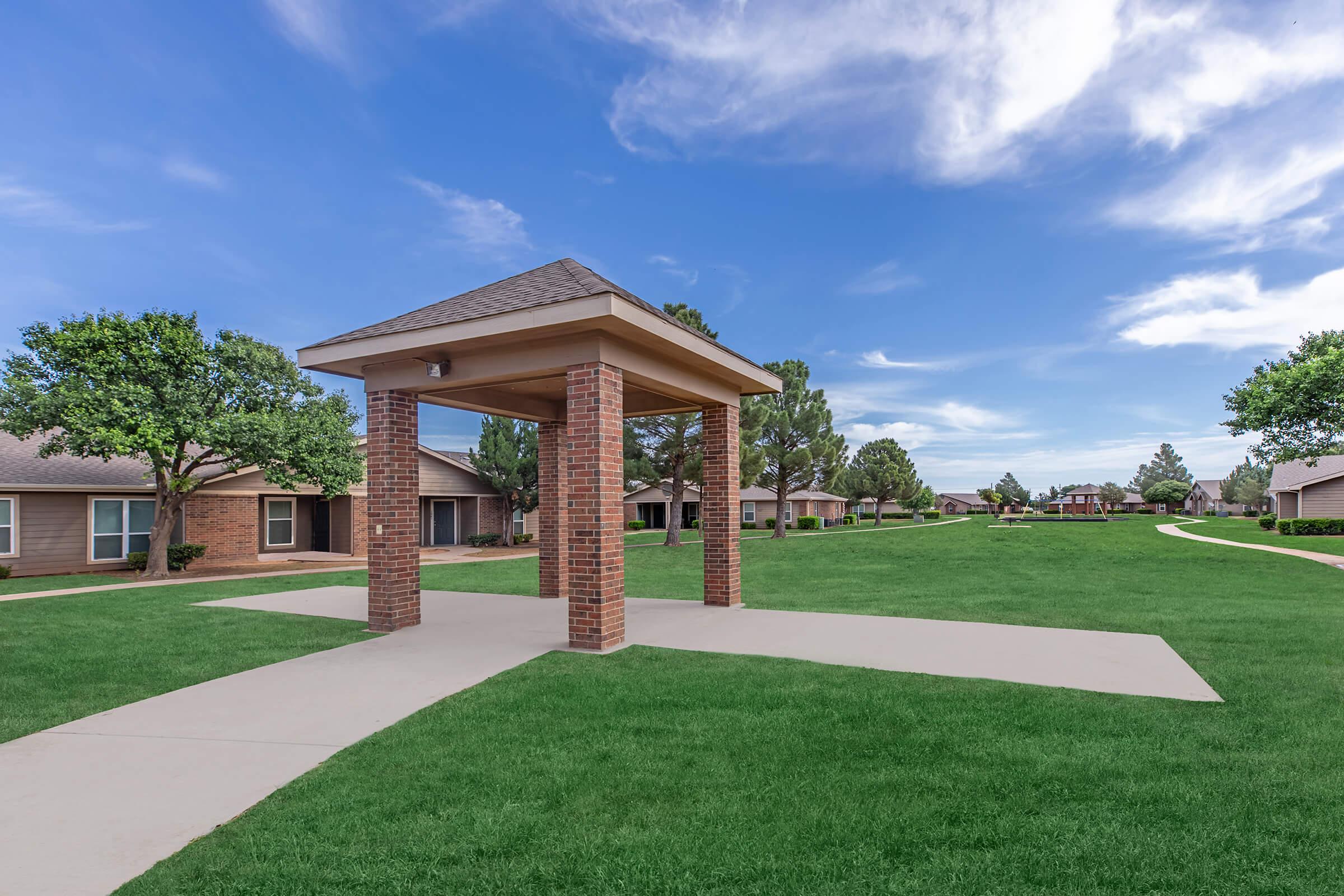 A landscaped area featuring a gazebo on a grassy lawn, surrounded by single-story homes. The sky is clear with a few clouds, creating a bright and inviting atmosphere. Pathways lead through the grass, and trees are visible in the background, enhancing the serene environment.