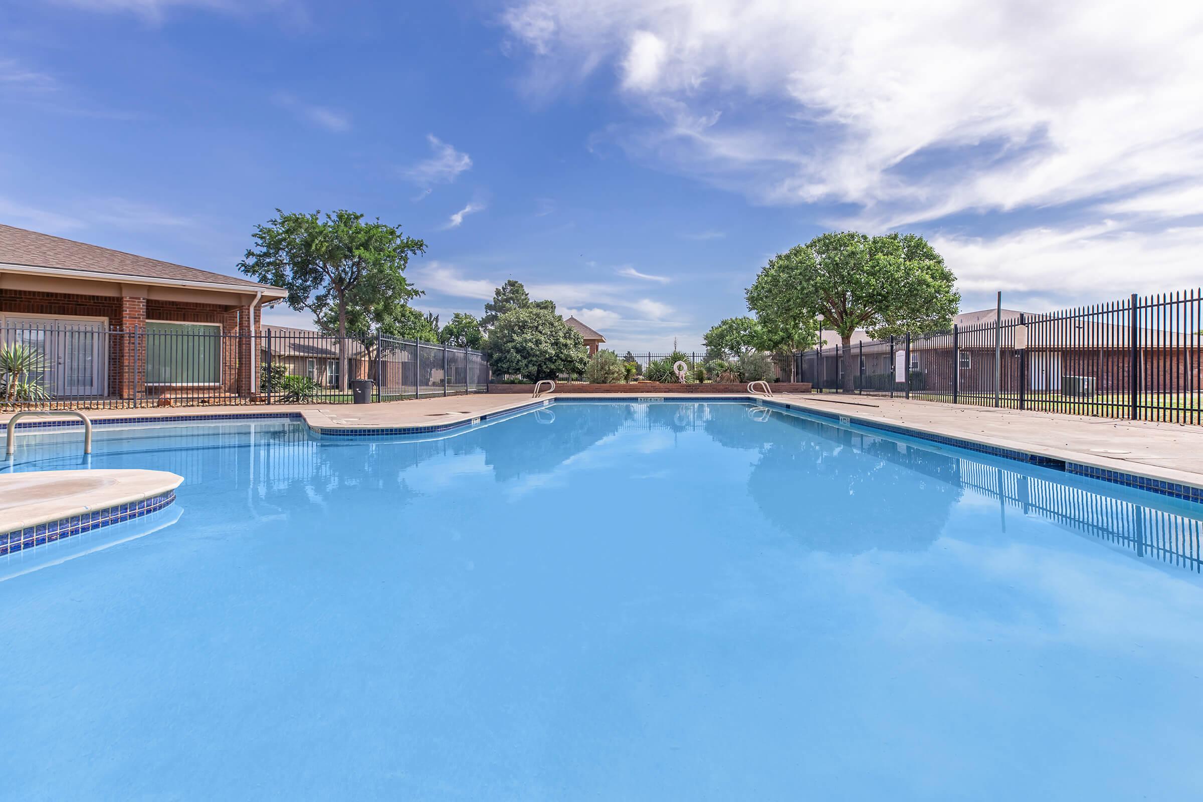 A clear blue swimming pool surrounded by a concrete deck, with lounge chairs and trees in the background. The sky above features white clouds and a bright blue hue, creating a sunny and inviting atmosphere. Fencing encloses the area, enhancing privacy and security.