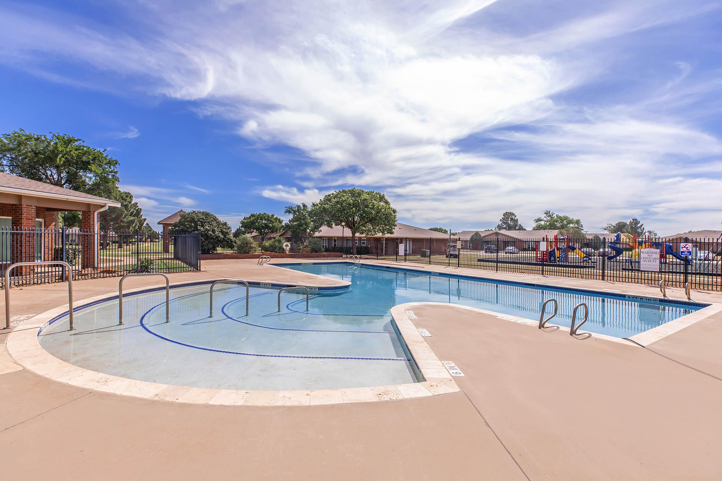 A spacious outdoor swimming pool with a curved design, surrounded by a concrete deck. In the background, there are residential buildings and trees under a bright blue sky with scattered clouds. Play structures are visible to the side of the pool area, indicating a family-friendly environment.