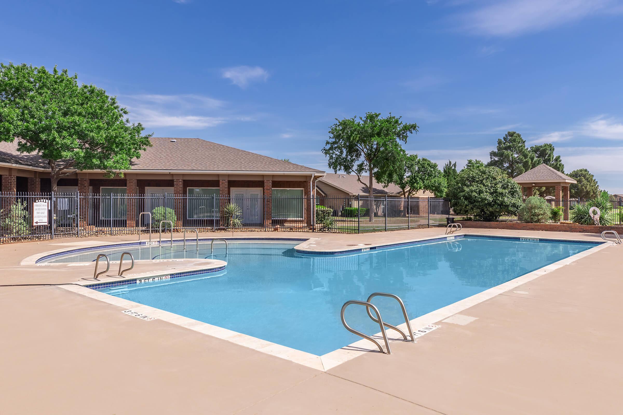Swimming pool surrounded by a paved area, featuring ladders for access. Green trees and shrubs frame the scene, with a residential building in the background. The sky is blue with a few clouds, and there is a fenced area around the pool for privacy.