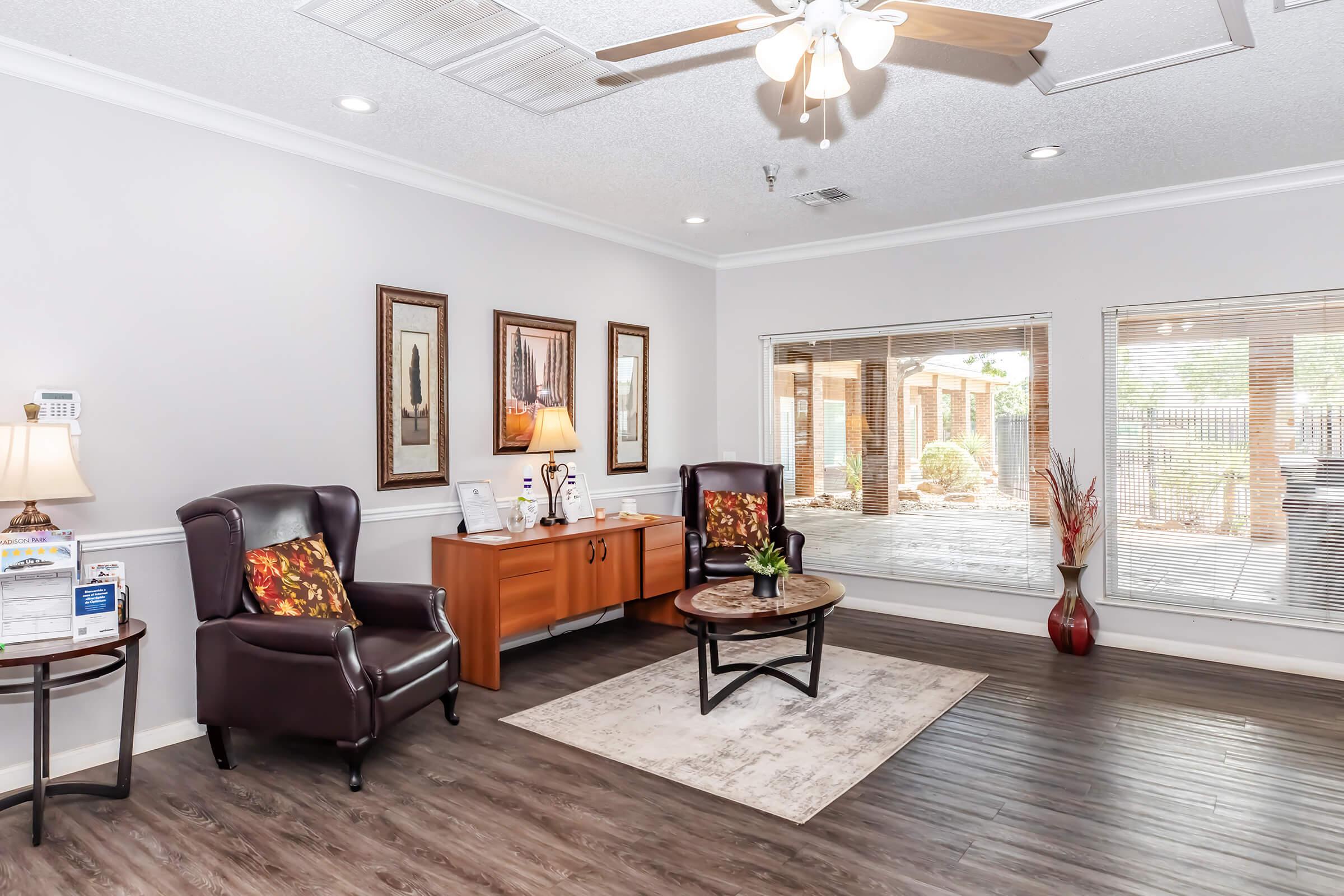 A cozy indoor sitting area featuring two dark leather armchairs with decorative pillows, a round coffee table, and a side table with a lamp. The walls are adorned with framed artwork, and a large window looks out to a patio with greenery. The flooring is dark wood, enhancing the inviting atmosphere.