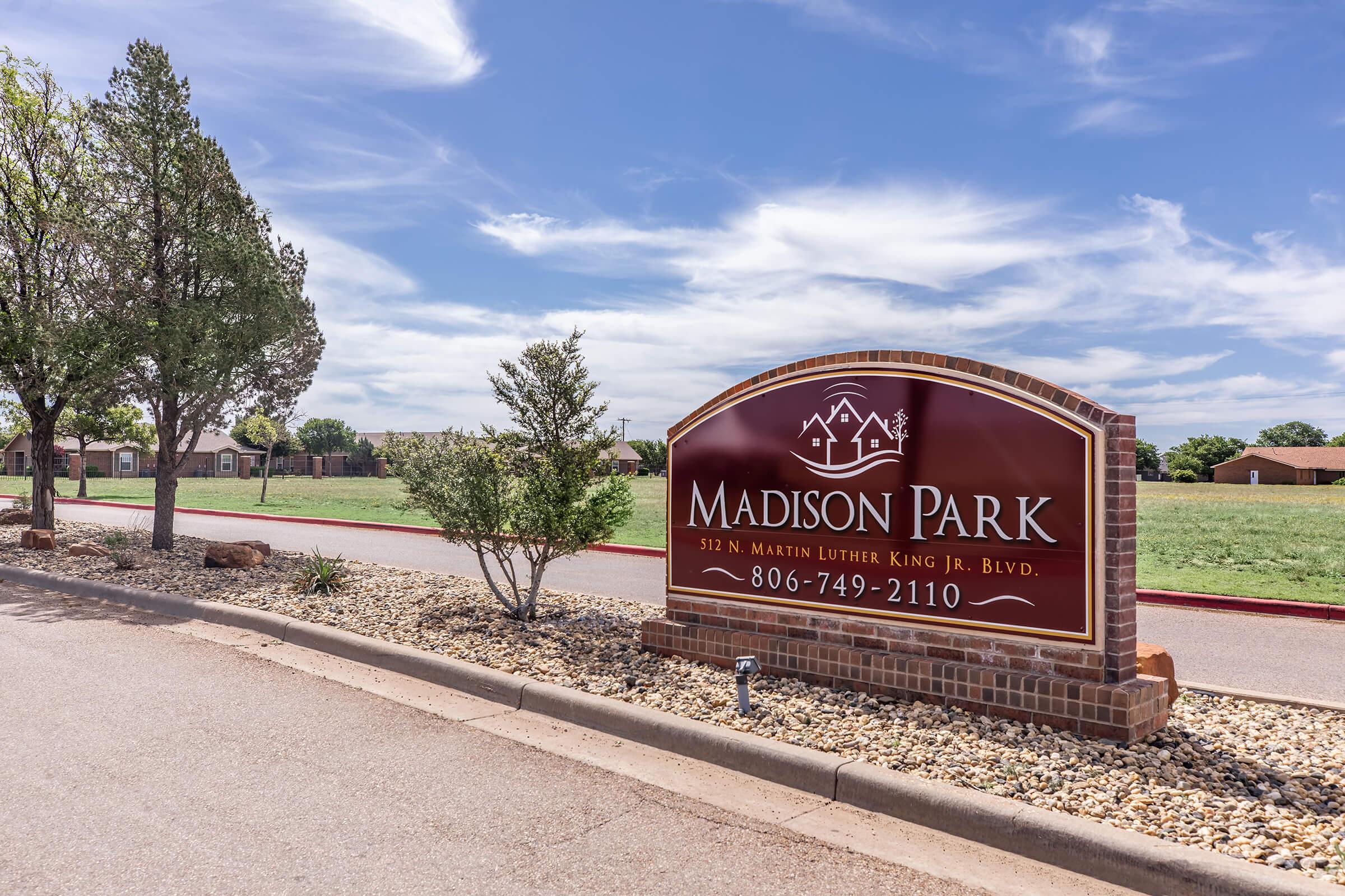 Sign for Madison Park located at 512 N. Martin Luther King Jr. Blvd. The sign features a brown background with white text, a logo, and a phone number, surrounded by landscaped greenery and a clear blue sky.