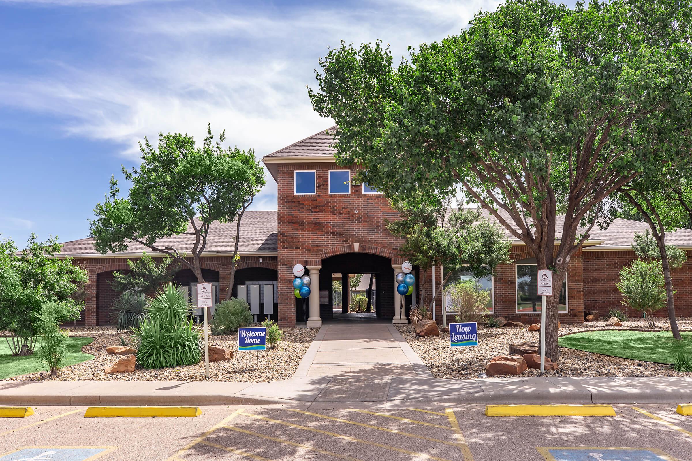 A welcoming brick building with a central entrance framed by green trees and landscaped stones. Signs on either side read "Welcome" and "Visit Us." The pathway leads to an open space, with blue and white balloons enhancing the inviting atmosphere. The sky is clear with a few clouds.