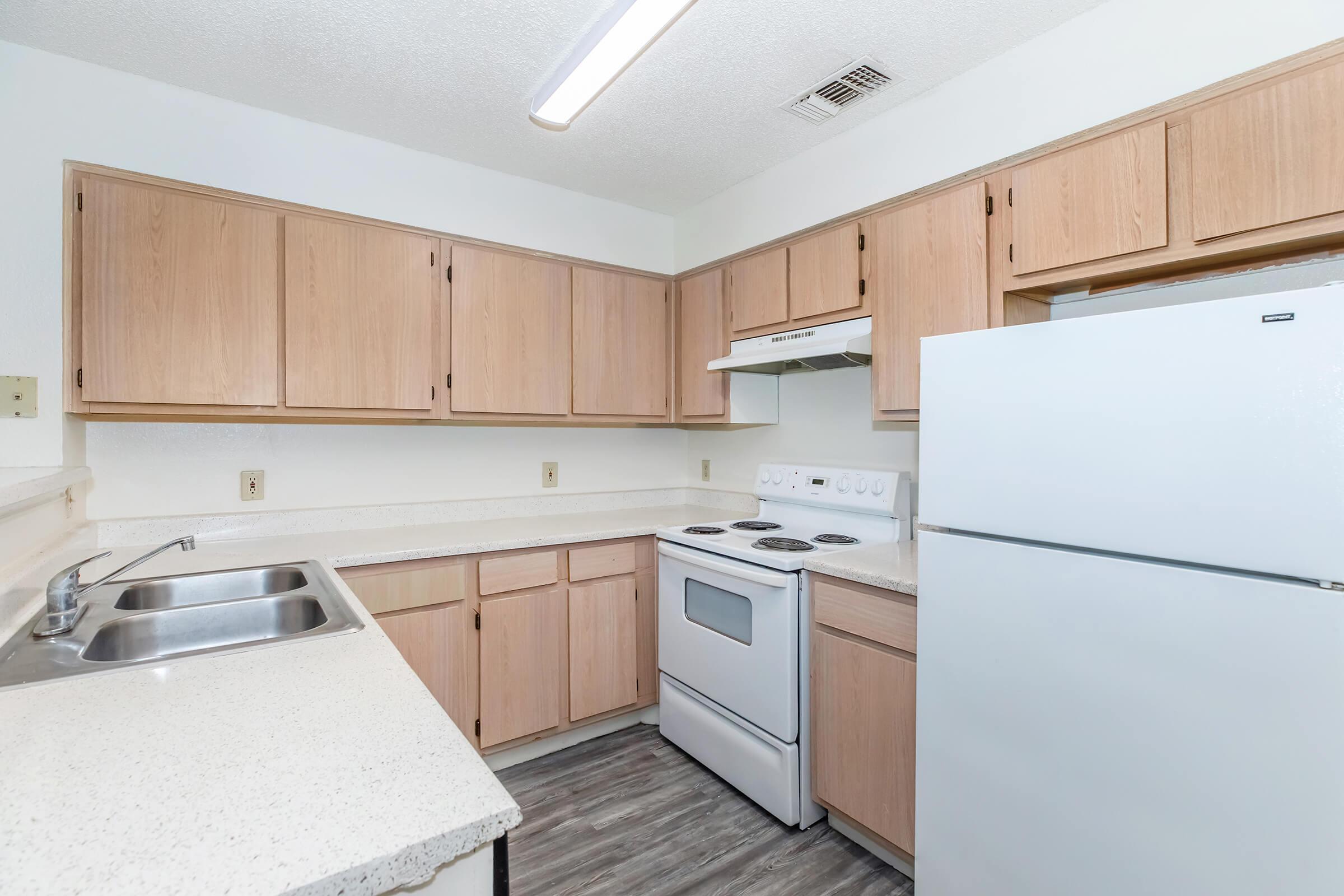 A clean and modern kitchen featuring light wood cabinets, a white stove and refrigerator, a double sink with a stainless steel basin, and a countertop. The walls are painted white, and there is a light fixture overhead. The flooring appears to be a wood-like laminate.