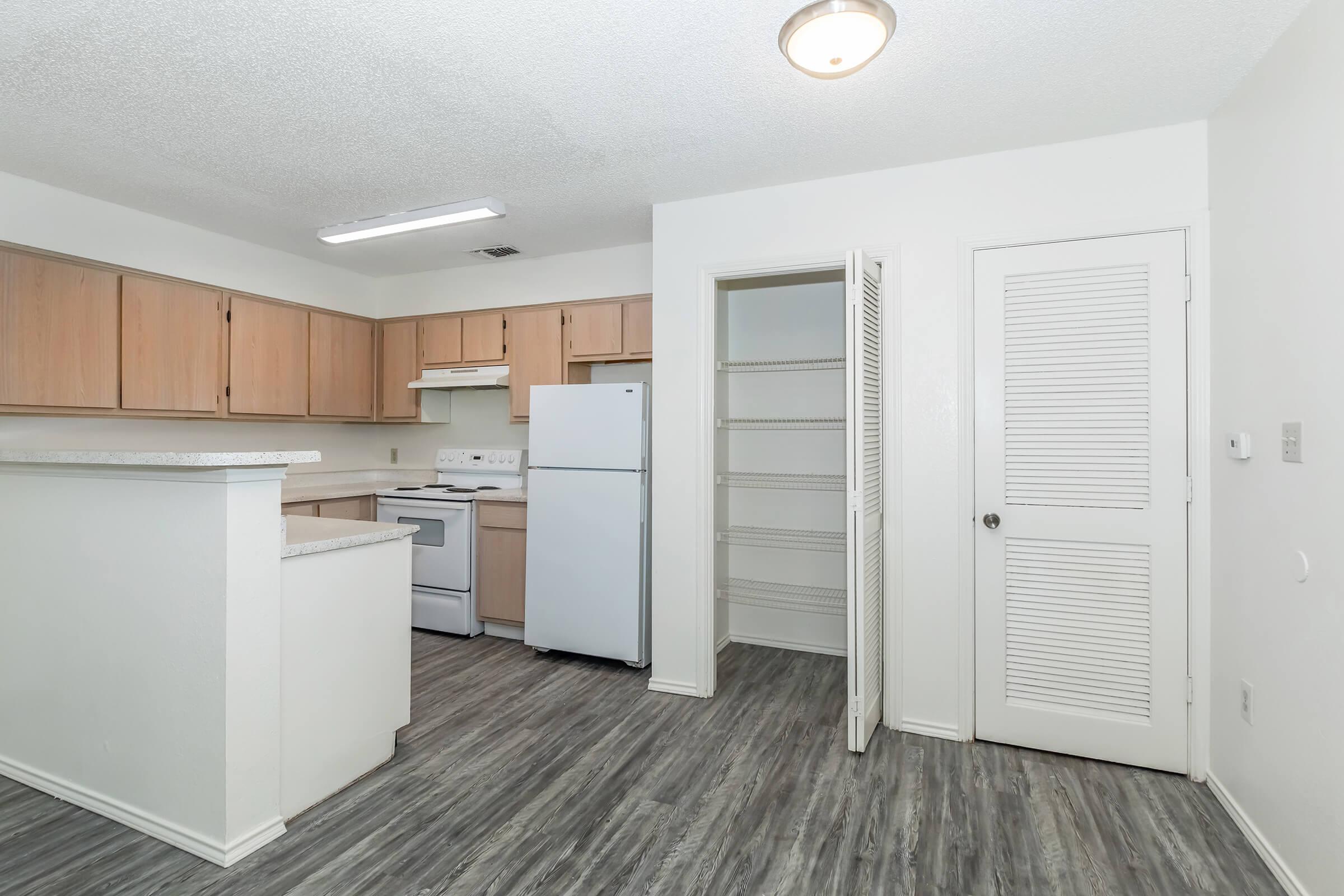 Spacious kitchen featuring wood cabinetry, stainless steel appliances including a refrigerator, and a pantry. The area has light-colored walls and laminate flooring, with a bright overhead light fixture.