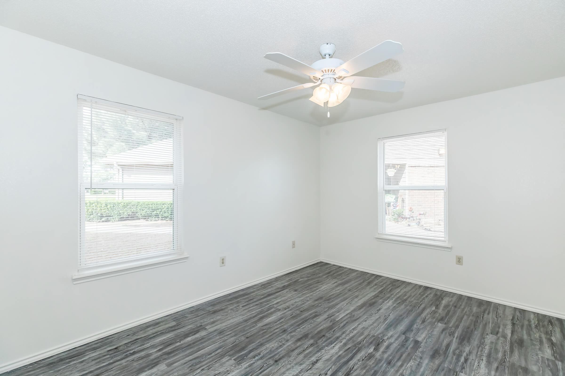 Empty room with light-colored walls and gray wood-like flooring. Two windows allow natural light to enter, and a ceiling fan with light fixture is centered in the room. The space is bright and uncluttered, creating a minimalist atmosphere.