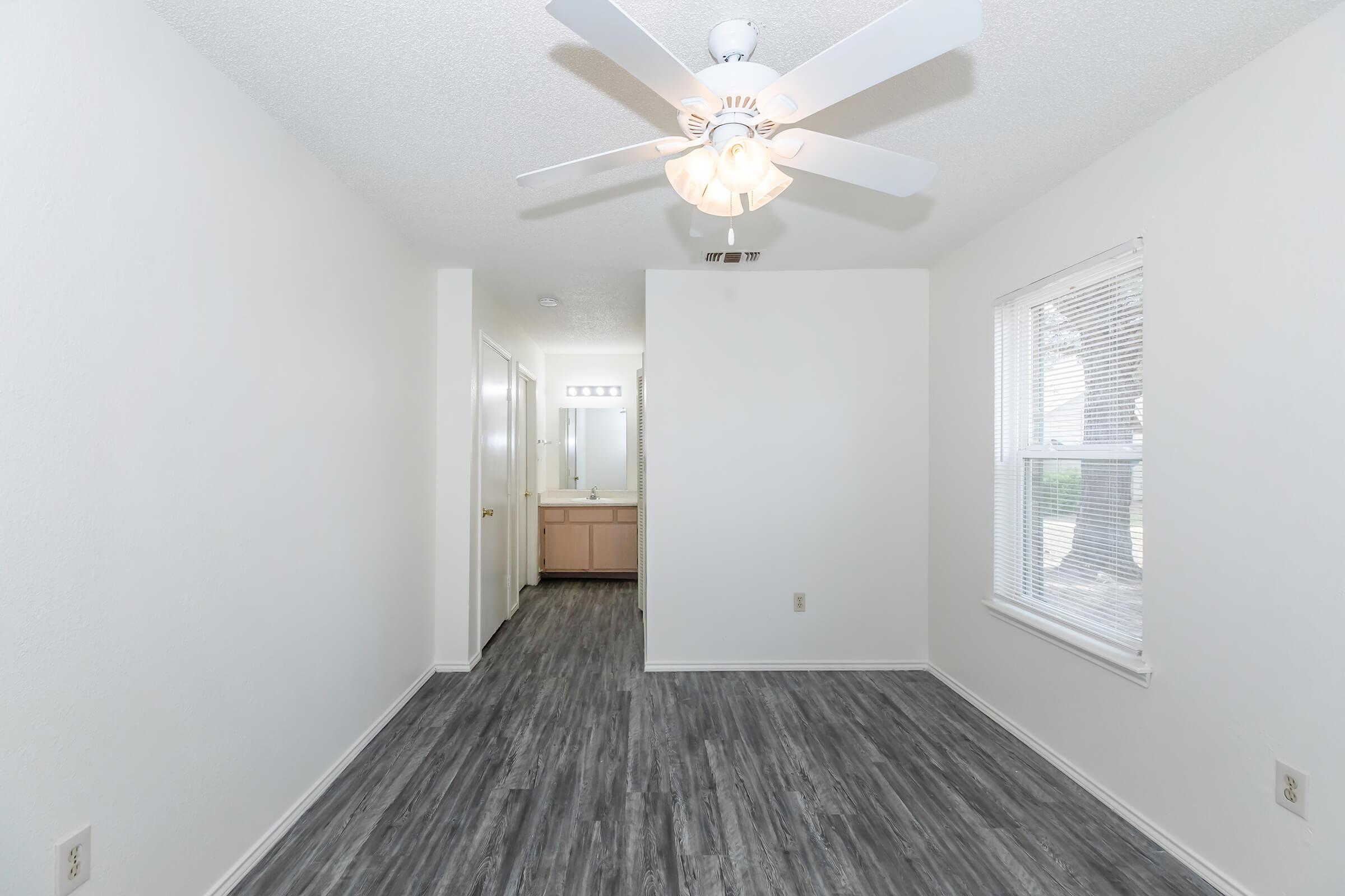 A bright, empty room featuring light gray laminate flooring, a ceiling fan with a light fixture, and white walls. A doorway leads to a bathroom with a vanity visible in the background. Natural light comes through the window, creating a spacious and airy atmosphere.