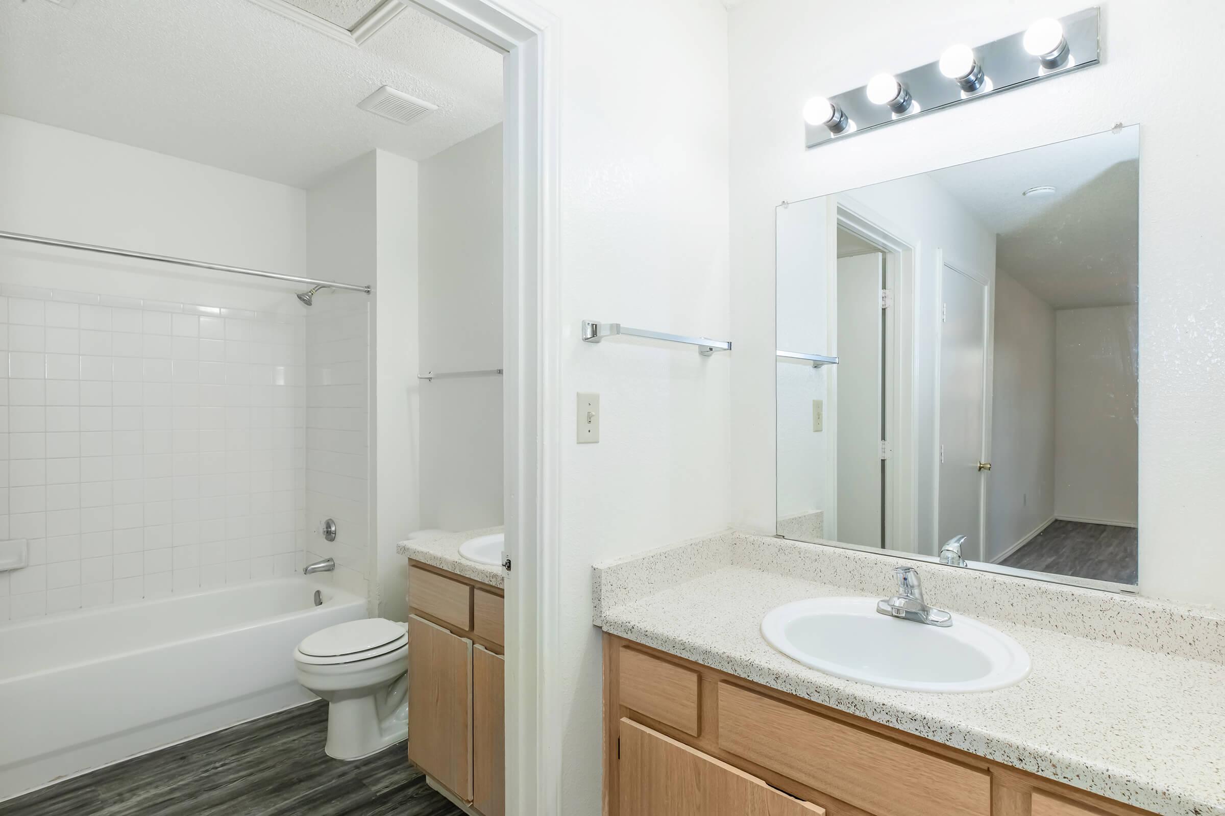 A clean and modern bathroom featuring a white bathtub with a shower, a toilet, and a single sink vanity with a lighted mirror. The walls are painted white, and the floor has a wood-like appearance. The room has a spacious feel with neutral colors throughout.