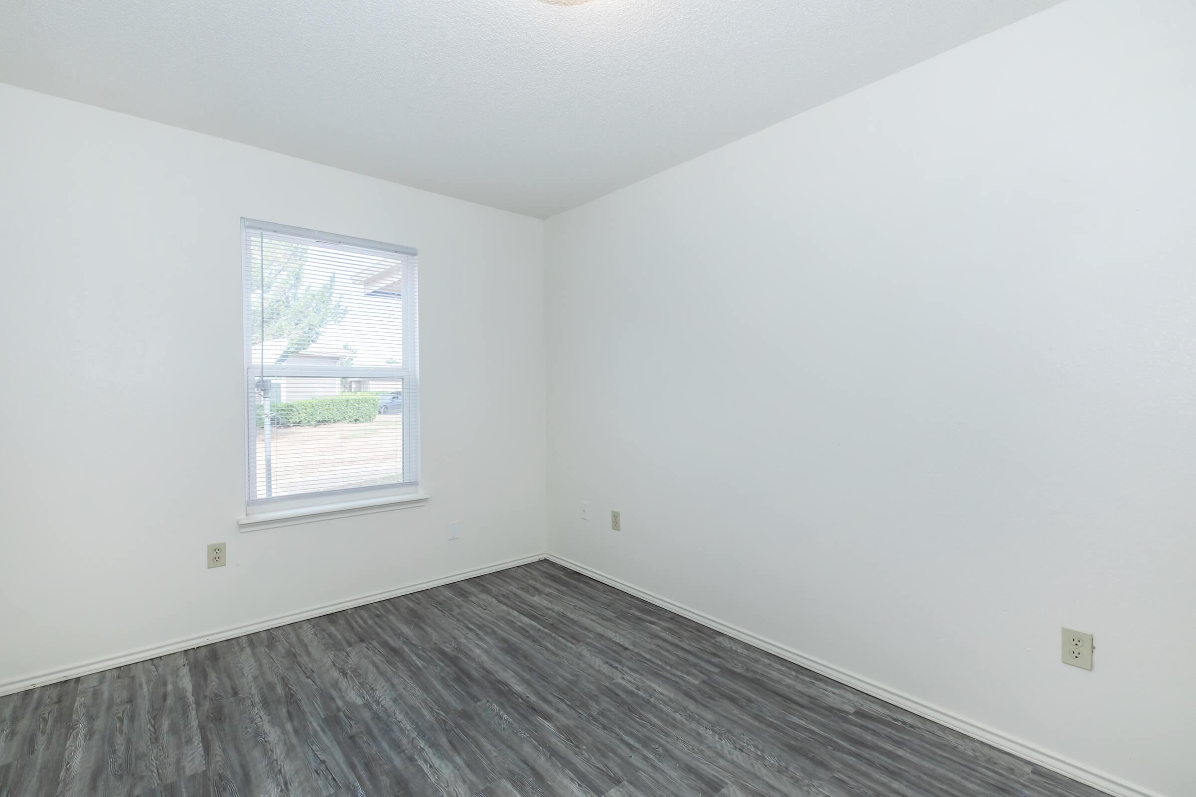 Empty room with a light-colored wall, a single window allowing natural light, and a dark wood-like floor. The space appears bright and minimalist, suitable for various uses or decoration.