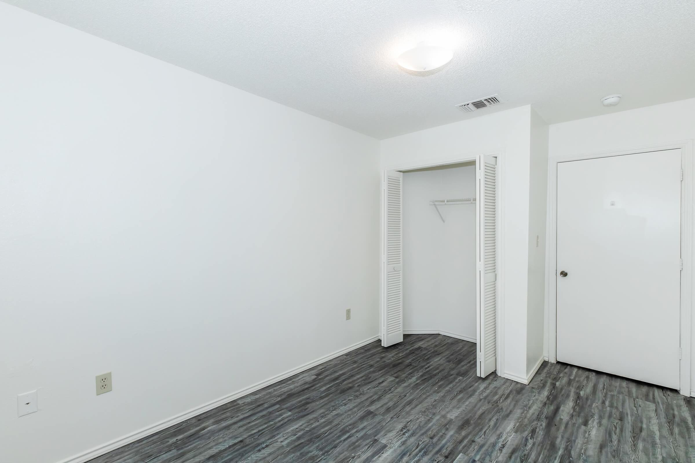 A vacant room with white walls and a ceiling light. The floor features dark laminate flooring. On the left, a closet with sliding doors is open, revealing a shelf inside. To the right, a closed door leads to another space. The overall atmosphere is minimalistic and bright.