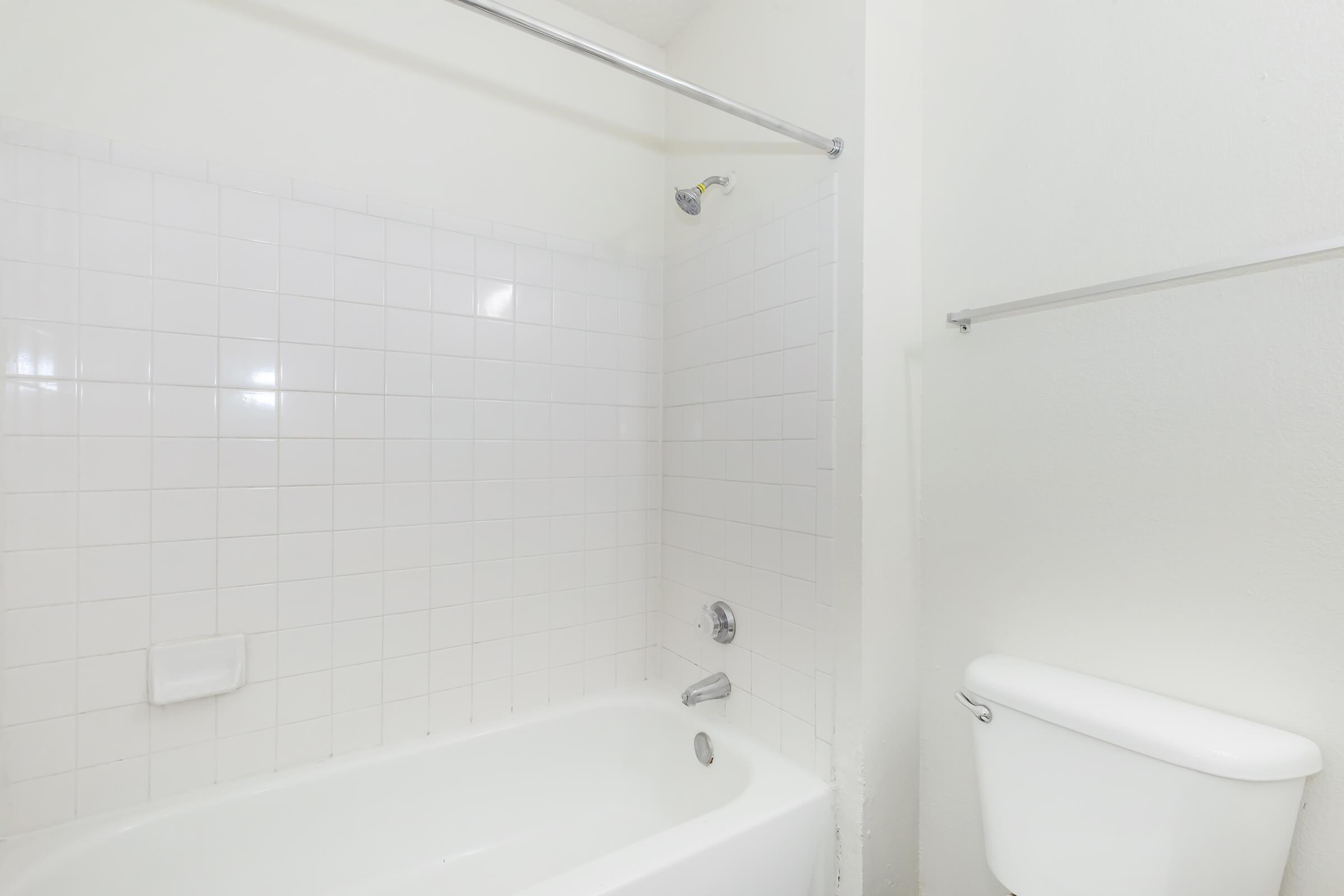 A clean, white bathroom featuring a bathtub with a showerhead above it. The walls are tiled with white square tiles, and a toilet is visible on the right side. The overall ambiance is bright and simple, with minimal decor.
