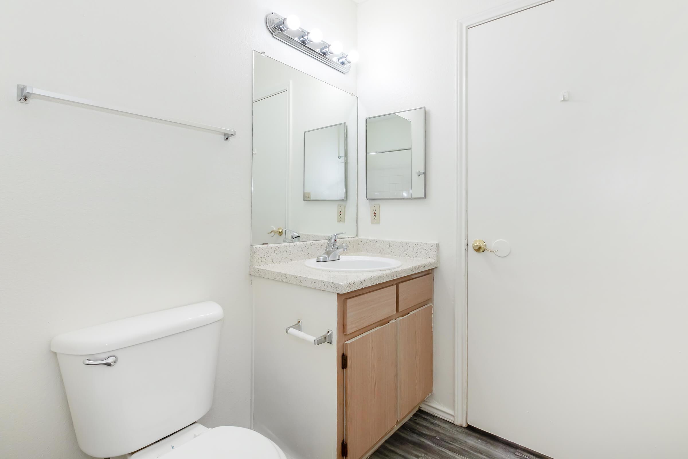 A small bathroom featuring a white toilet, a sink with a granite countertop, and a large mirror above the sink. There are two additional smaller mirrors on the wall, and the room has white walls and a light fixture above the mirror. A closed door is visible on the right side of the image.