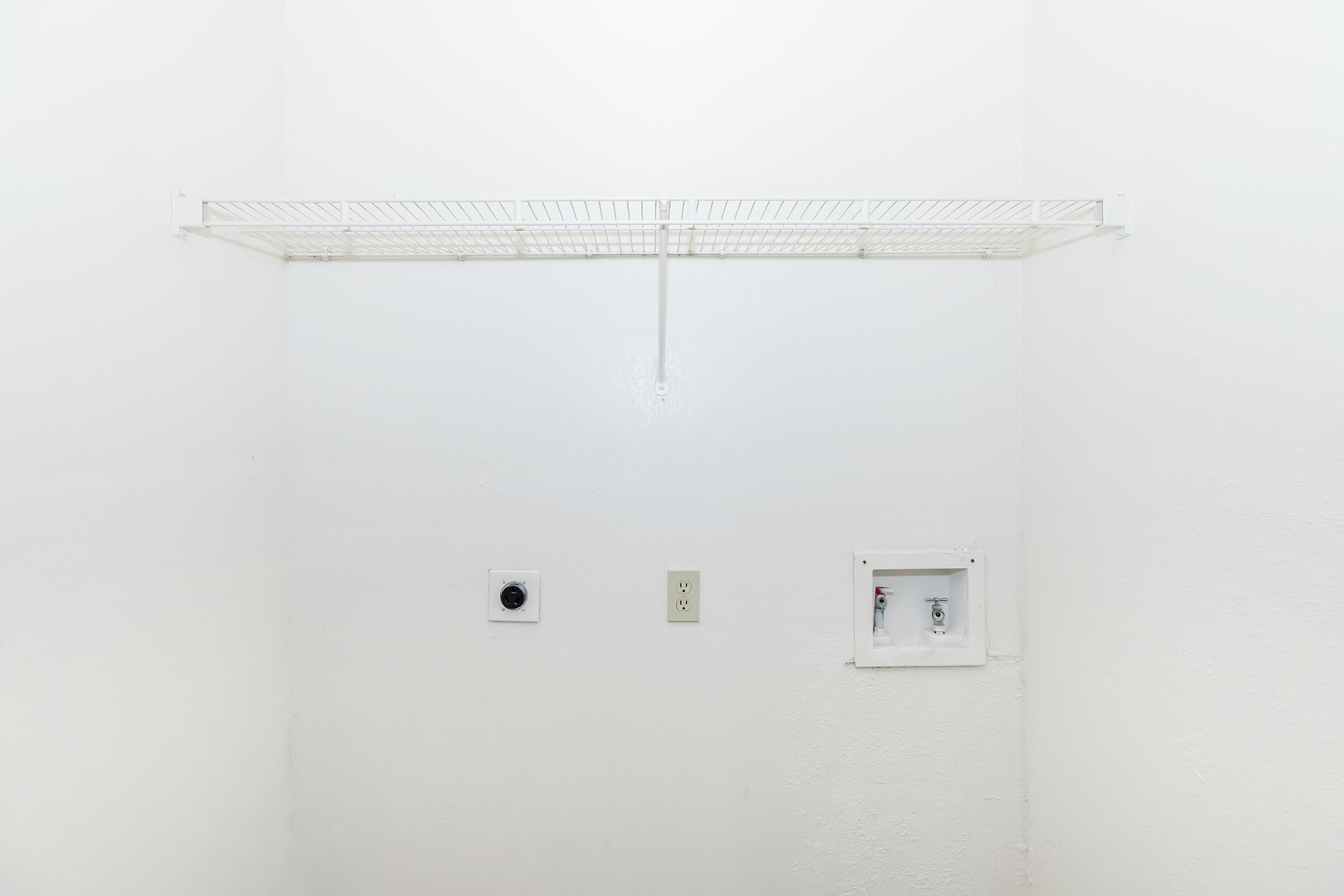 A minimalistic laundry area with a single white wire shelf mounted on a plain white wall. To the left, there's a wall outlet and a round, black electrical socket, while a small, rectangular panel is visible to the right, possibly for plumbing access. The space appears clean and uncluttered.
