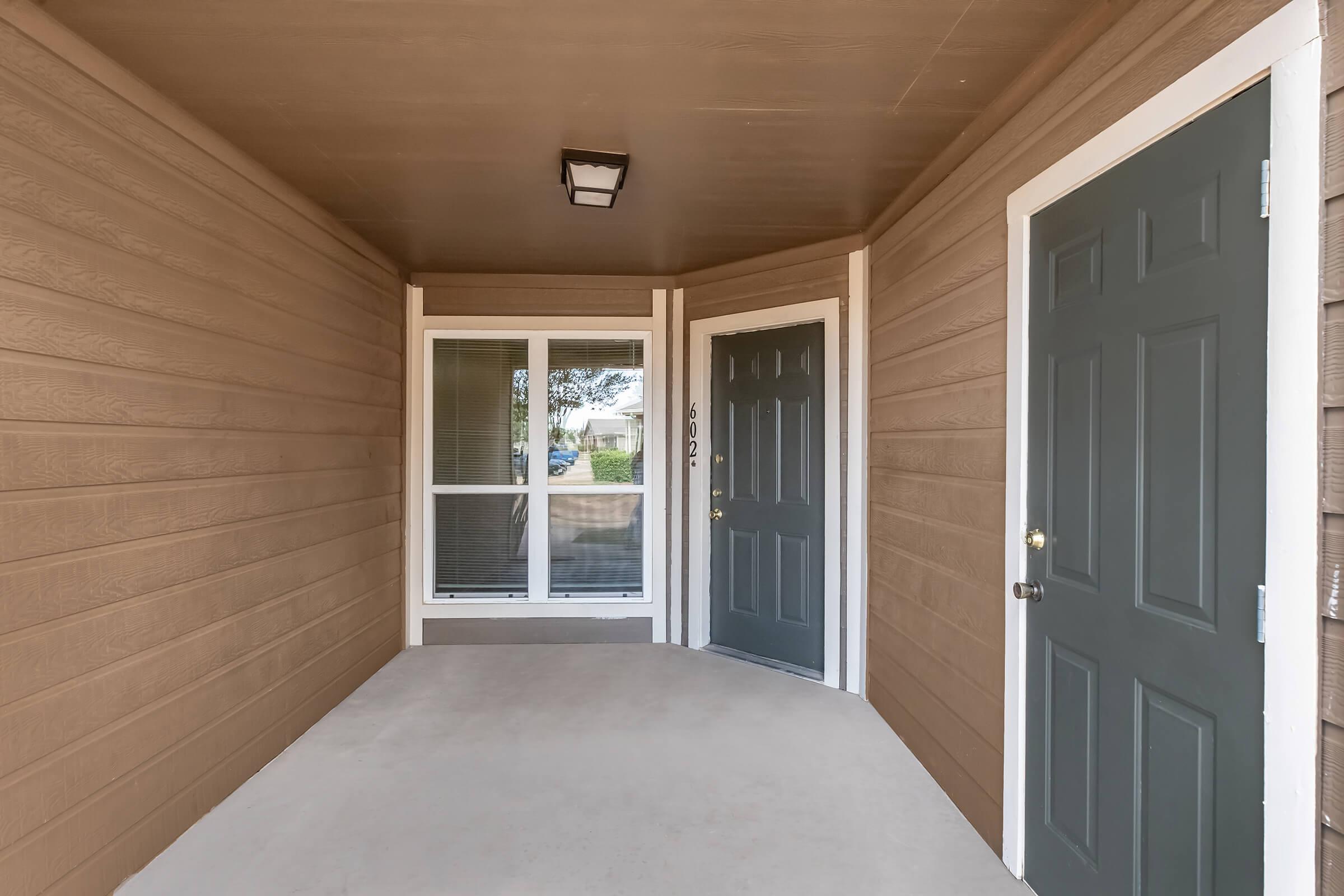 A covered porch area featuring a smooth concrete floor and two dark green doors. One door is a standard entry door, while the other is a windowed door leading to the interior. The walls are finished in light brown horizontal siding, and there is a ceiling light fixture. A view of the outside can be seen through the window.