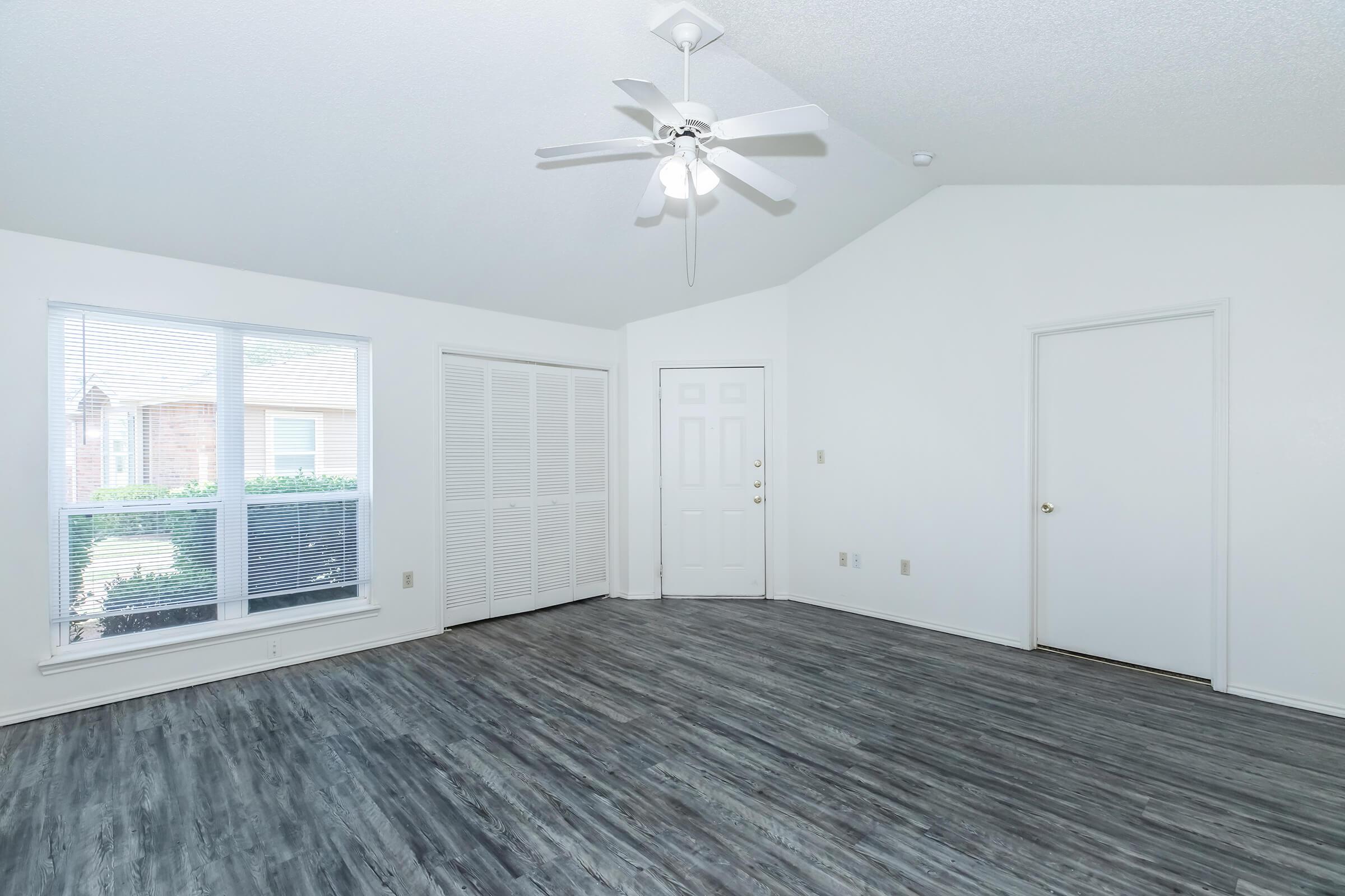 A bright, empty room with a vaulted ceiling, featuring a ceiling fan. Large windows provide natural light, and there are white walls and modern, dark wood-like flooring. A closet with sliding doors is visible, along with a door leading outside.