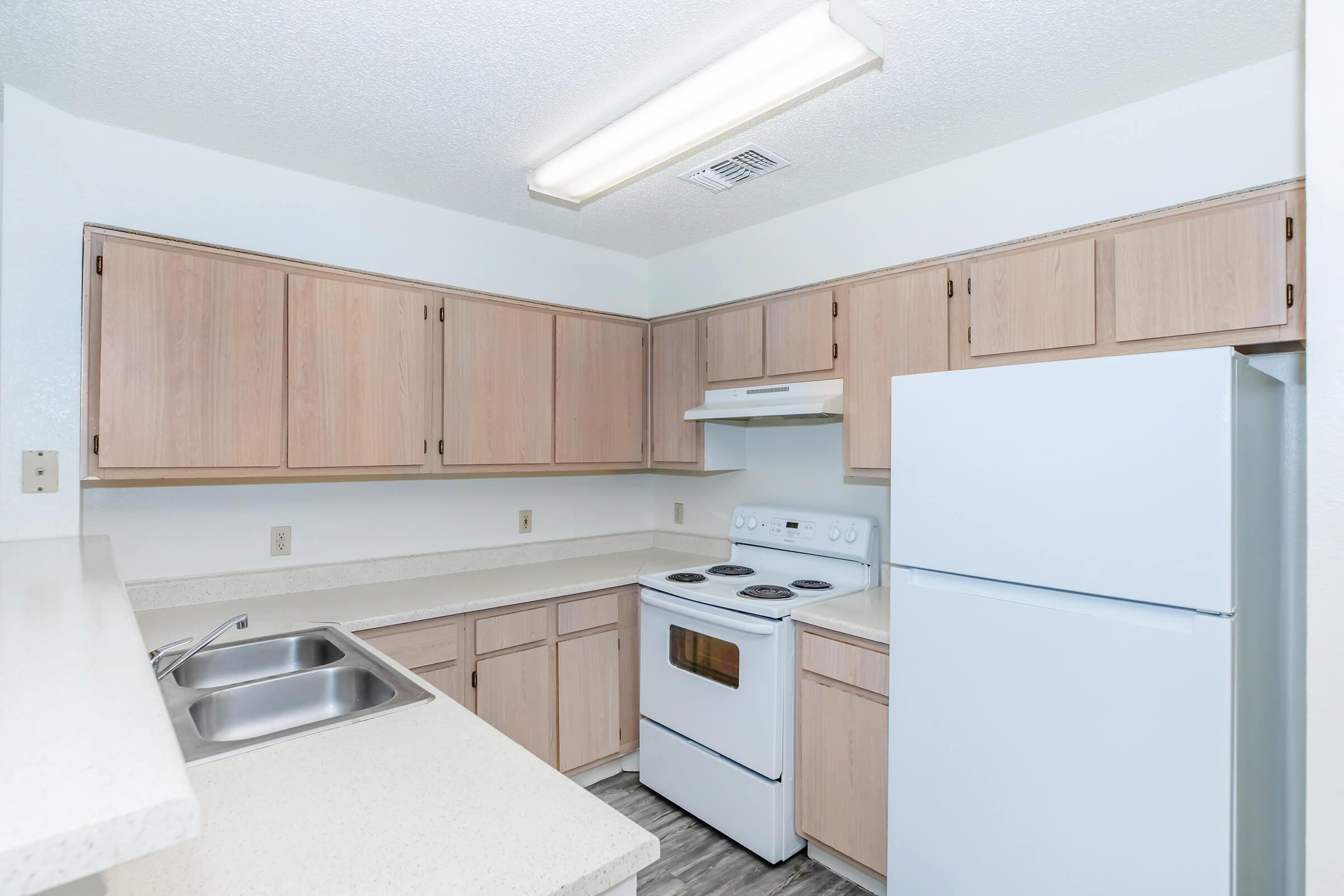 Bright kitchen featuring light-colored wooden cabinets, a white refrigerator, and an electric stove. The counter is made of a light-colored material, and there's a stainless steel sink. The walls are painted white, creating an airy and open atmosphere.