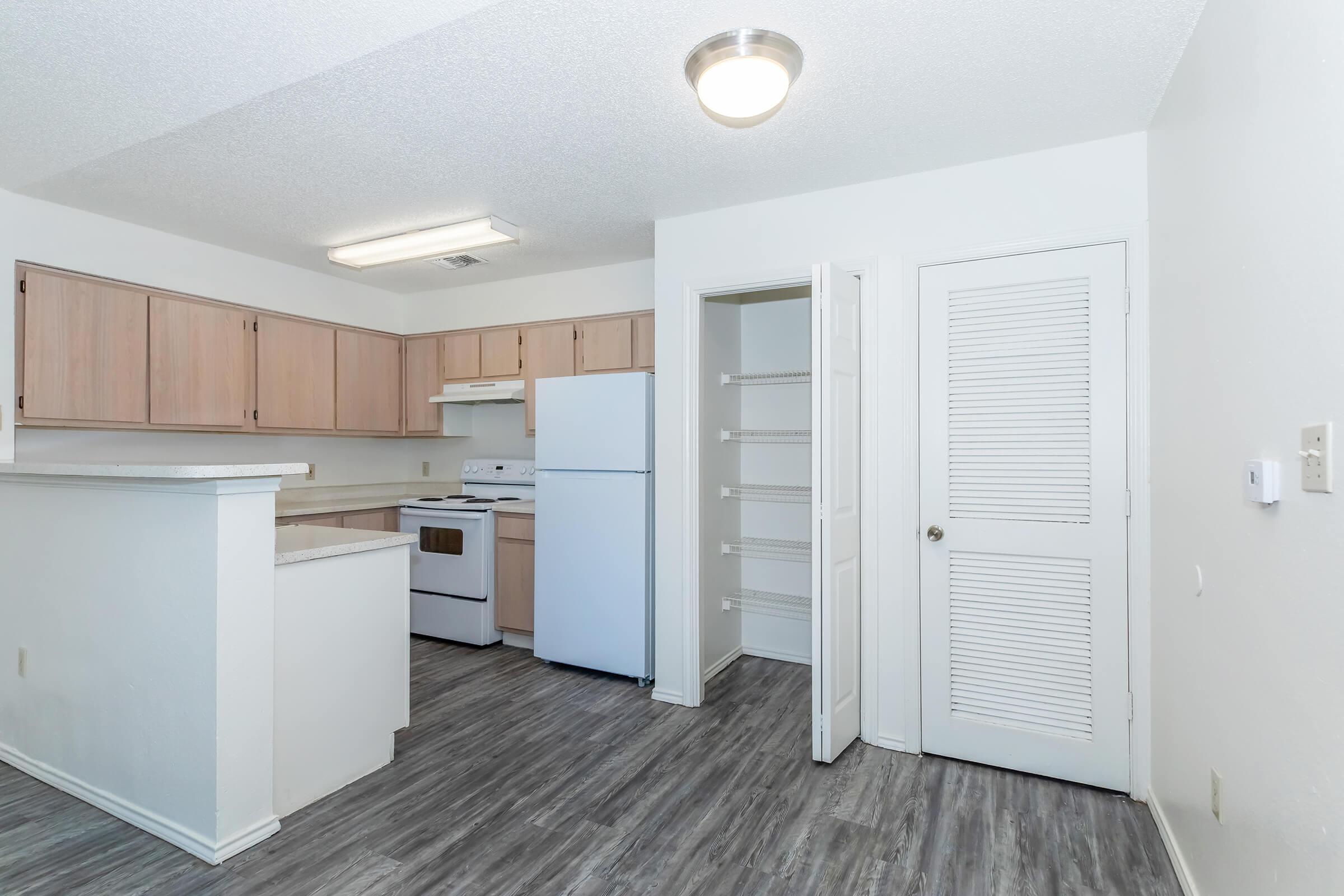A bright and modern kitchen with light wood cabinets, a white stove and refrigerator, and gray laminate flooring. A pantry with sliding doors is visible next to the entrance. The walls are painted in a light color, creating an open and airy atmosphere.