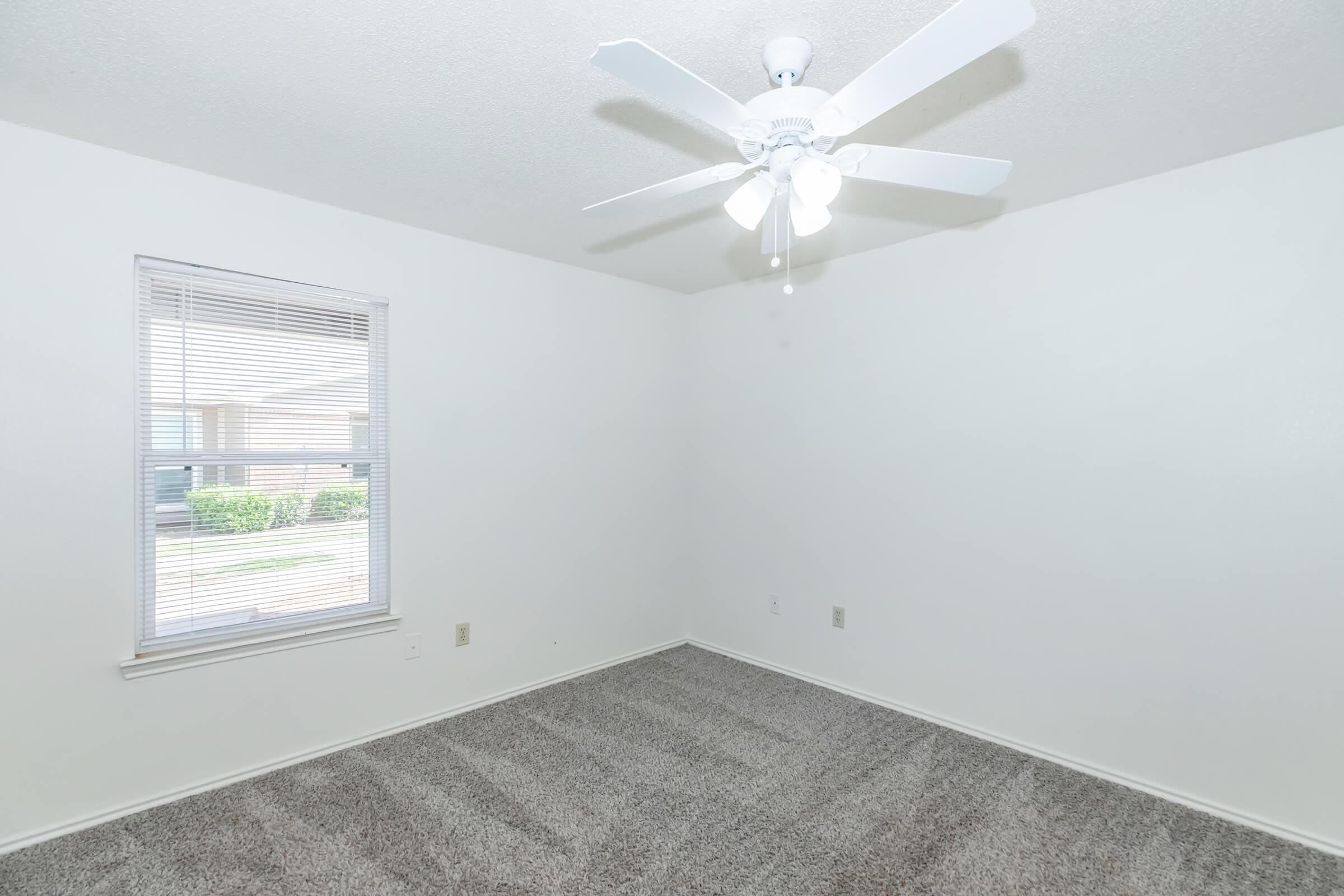 A vacant room with light gray carpet and white walls. A ceiling fan with bright lights is centered in the ceiling. There's a window with blinds on the left side, allowing natural light to enter. The space is clean and unfurnished, creating an open and airy feel.