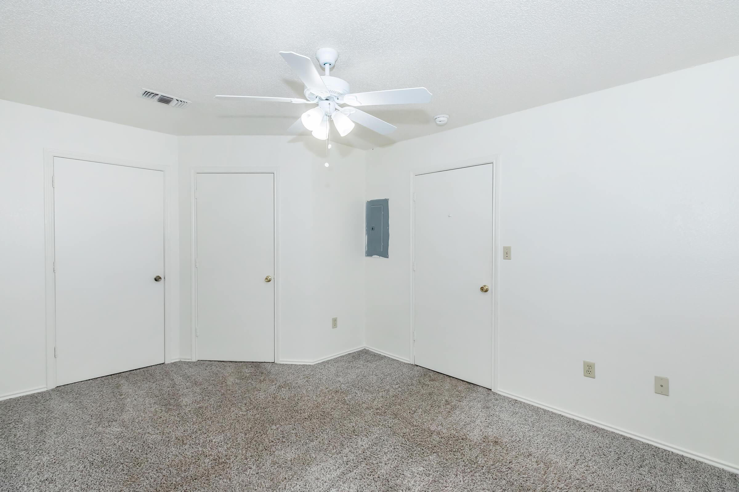 A spacious, empty room with light beige carpet and white walls. A ceiling fan with five blades hangs from the ceiling, providing light. There are two closed doors on one wall and an electrical panel mounted on another wall. The room is well-lit and bright, featuring a simple, clean design.