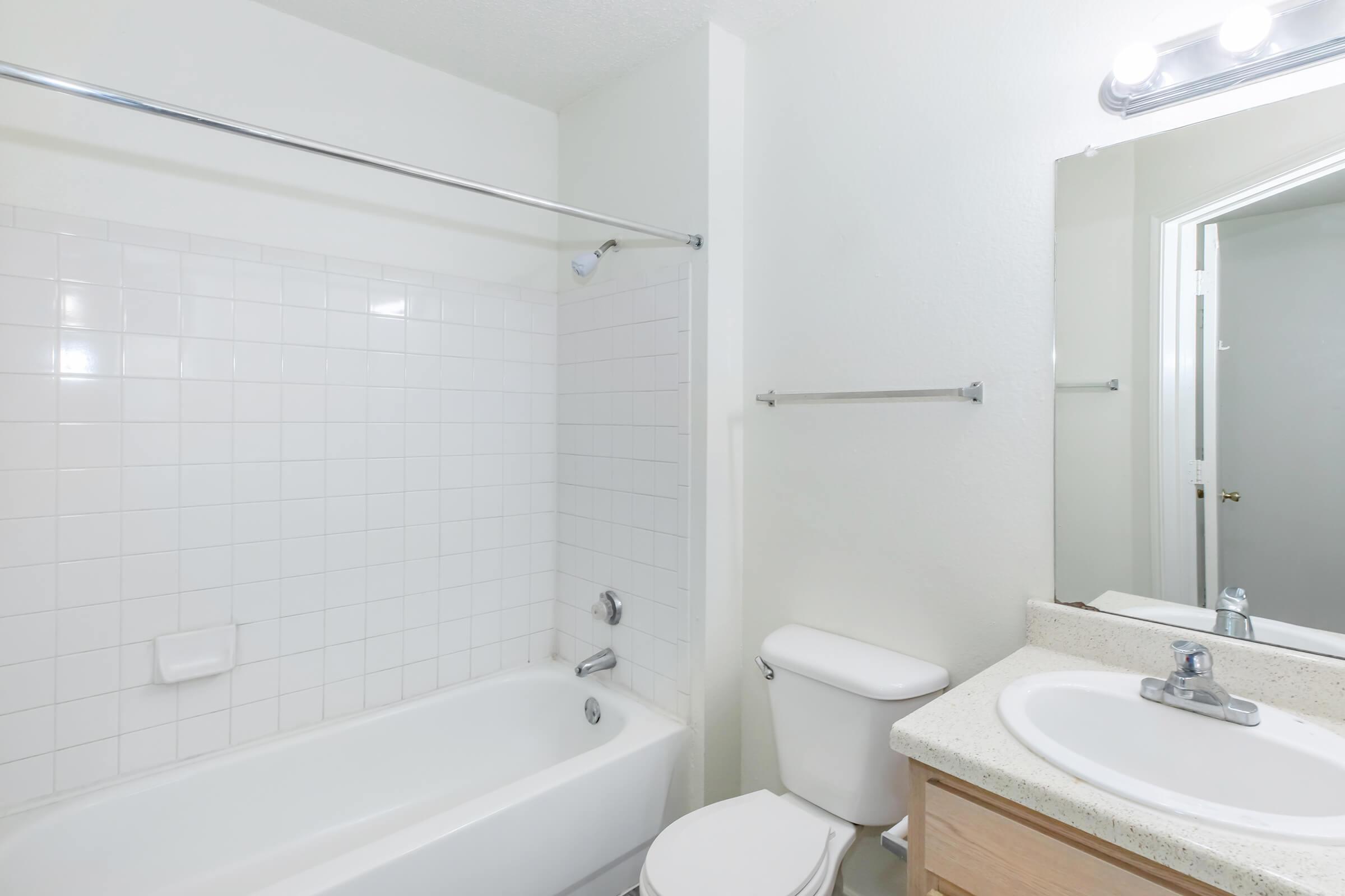A clean and simple bathroom featuring a white tiled shower/bathtub, a toilet, and a sink with a countertop. The walls are painted white, and there is a mirror above the sink with a light fixture above it. The overall design is minimalist and tidy.