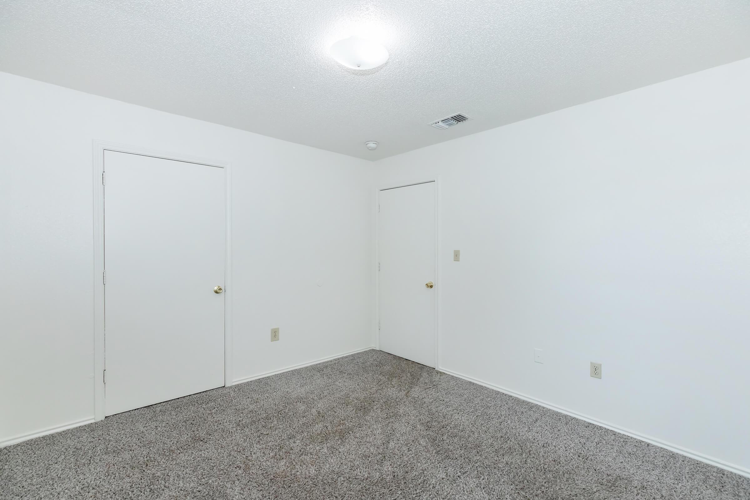 A vacant room with light-colored walls and a textured ceiling. The floor is covered with gray carpet. Two white doors are visible, one on the left and another on the right, leading to different spaces. The room is brightly lit by a central ceiling light fixture.