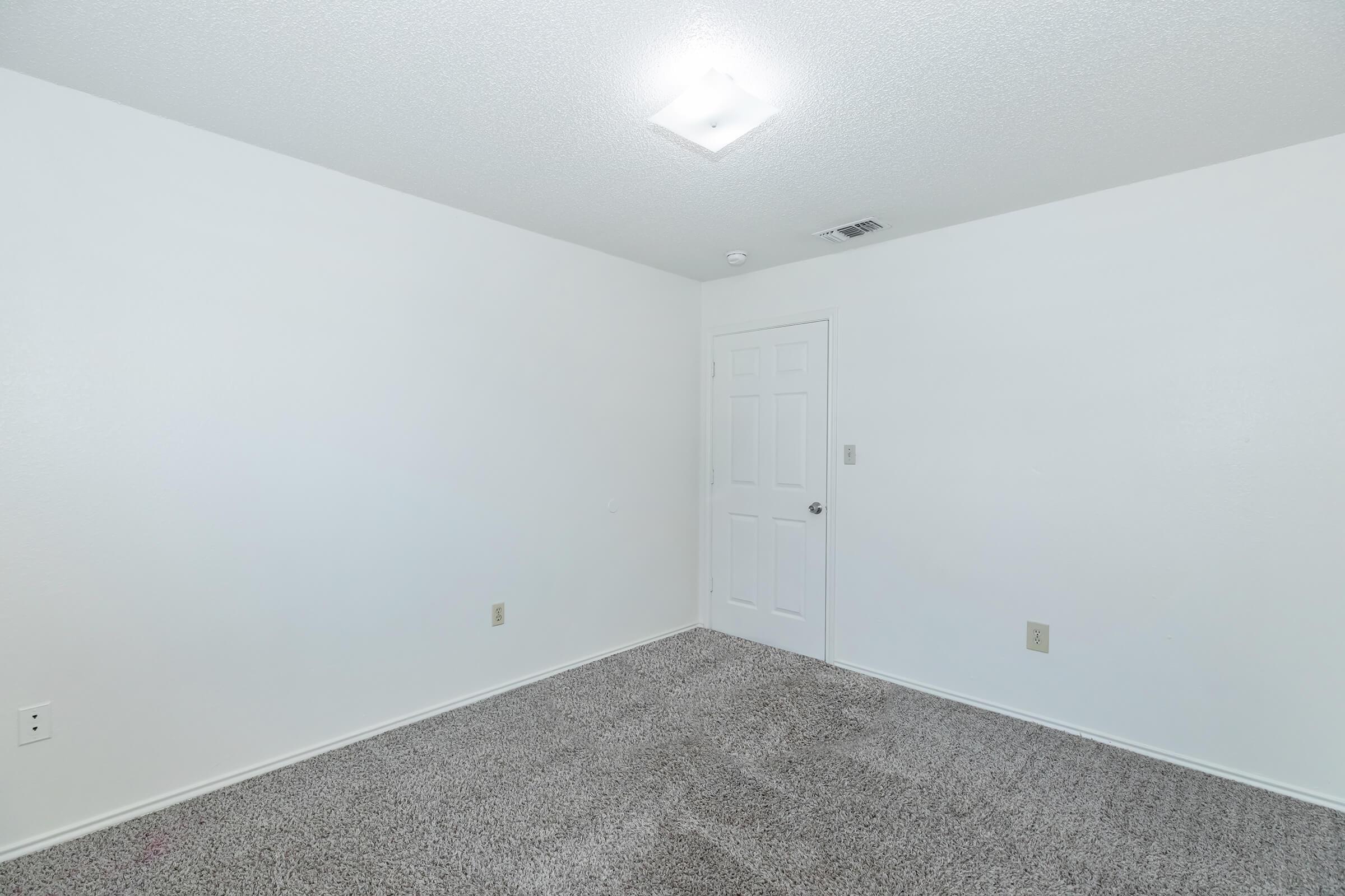 A vacant room featuring light-colored walls and carpet. The ceiling has a square light fixture, and there is a closed white door on one wall. The space is empty, giving it a clean and simple appearance.