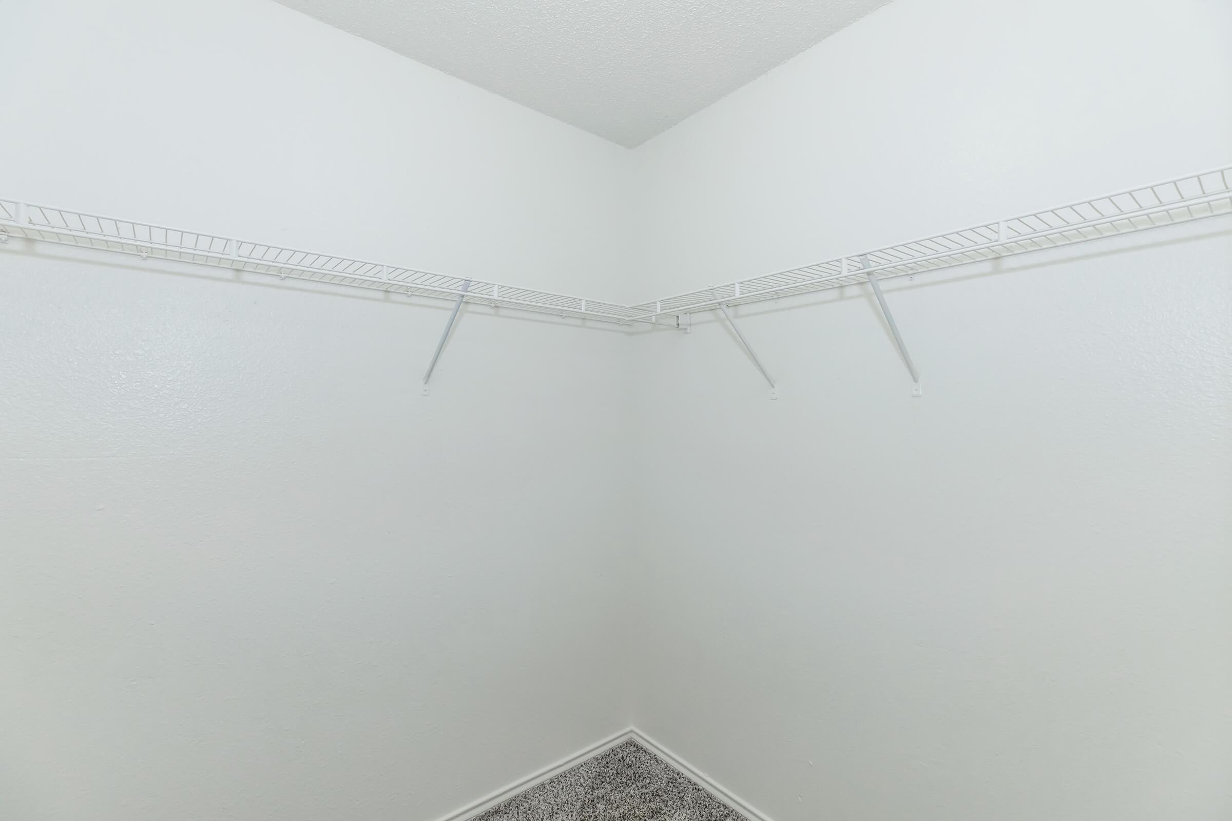 Empty corner closet with white walls and two wire shelves. The floor features a textured black and white surface.