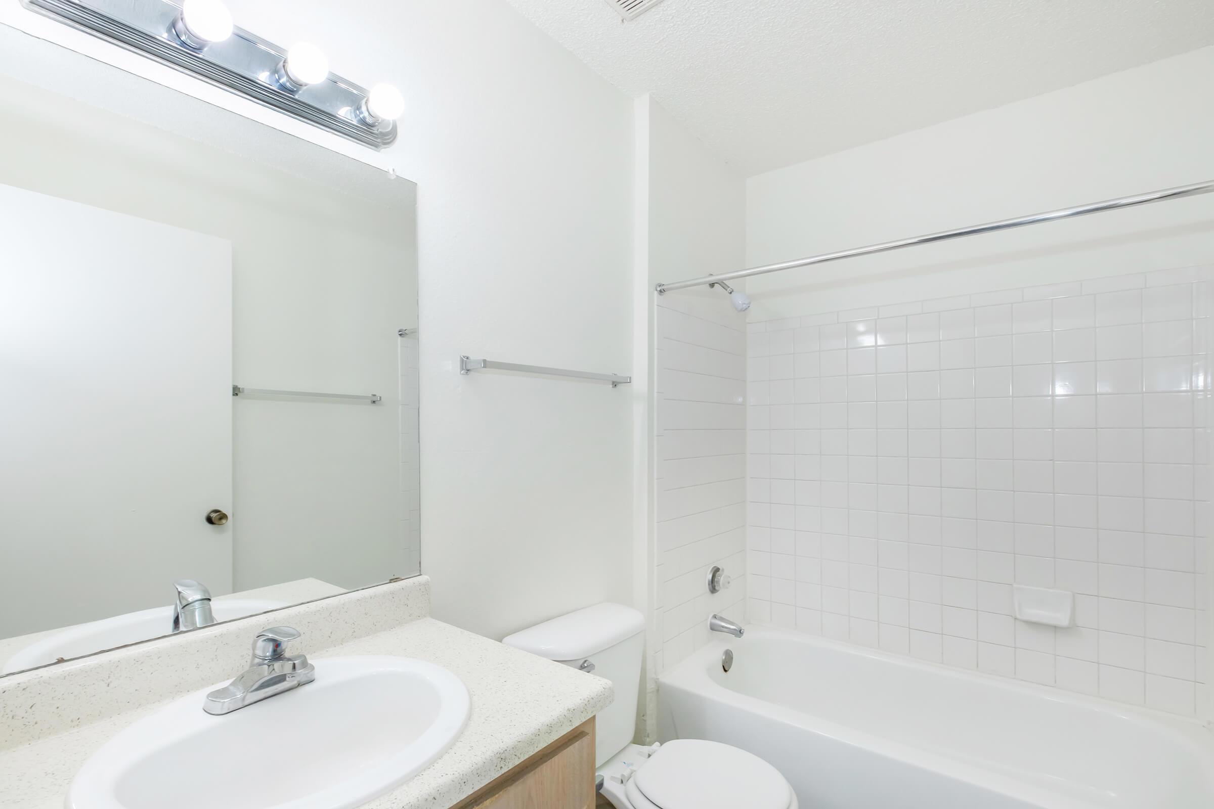 A clean, bright bathroom with a white tiled shower and bathtub. The vanity has a round sink with a chrome faucet, a light fixture above a mirror, and a simple countertop. The walls are painted white, and there is a towel rack mounted on one side. The atmosphere is fresh and modern.