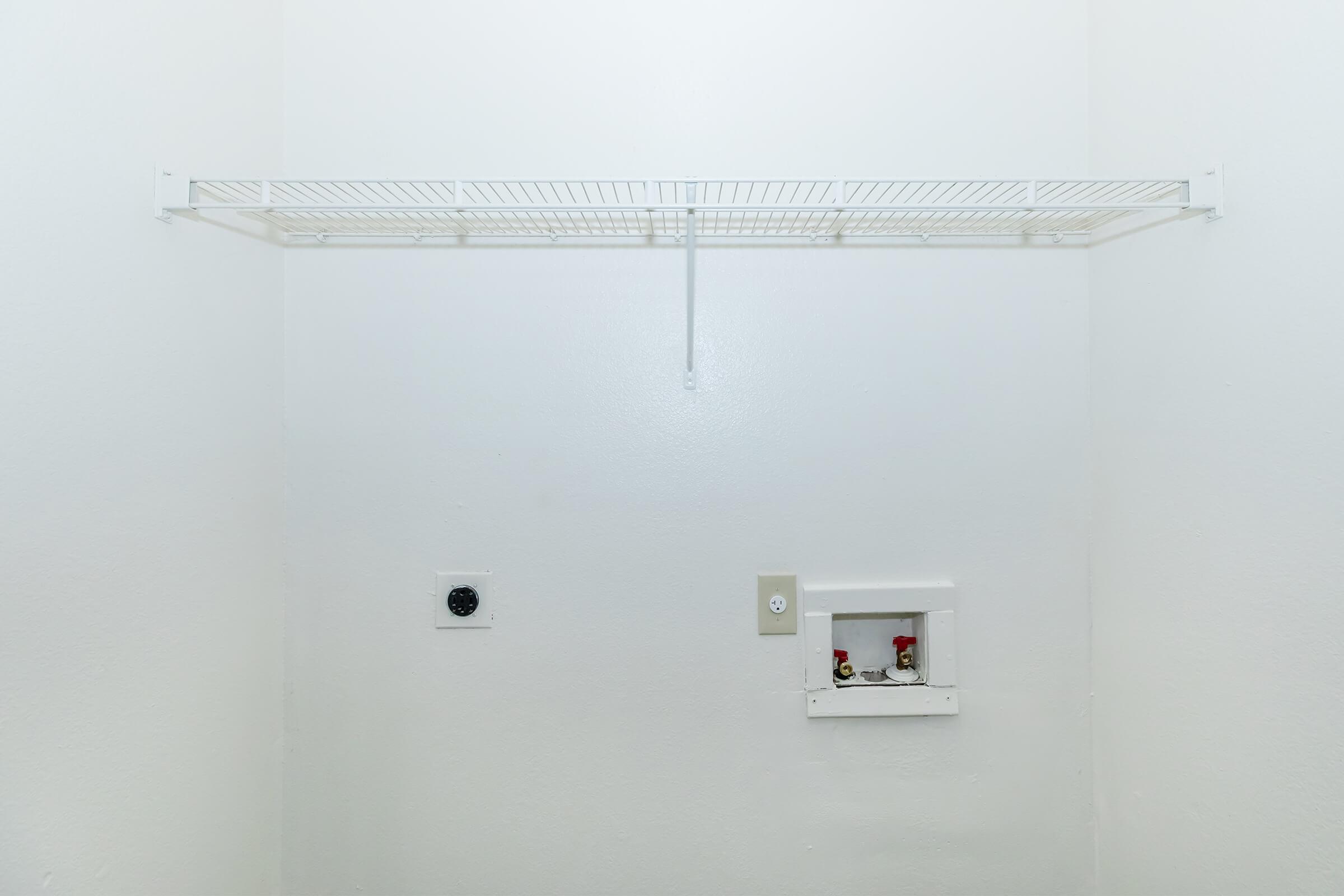 A simple, empty room with white walls featuring a wire shelving unit mounted on one side. There are two electrical outlets and a small panel in the wall, possibly for plumbing or utilities. The overall appearance is minimalistic and uncluttered.