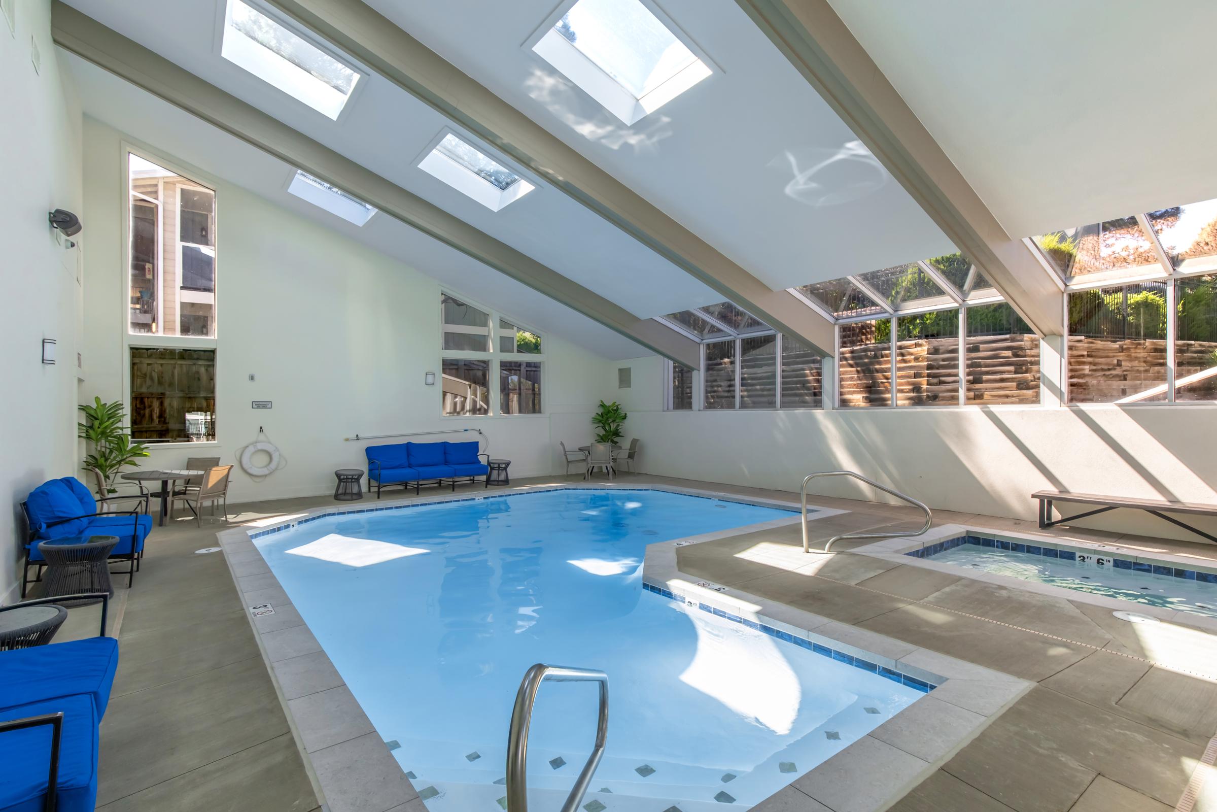 Indoor swimming pool area featuring a rectangular pool with a spa, surrounded by large windows that allow natural light in. The space includes blue lounge chairs and a structured ceiling with skylights. The atmosphere is bright and inviting, ideal for relaxation and leisure activities.