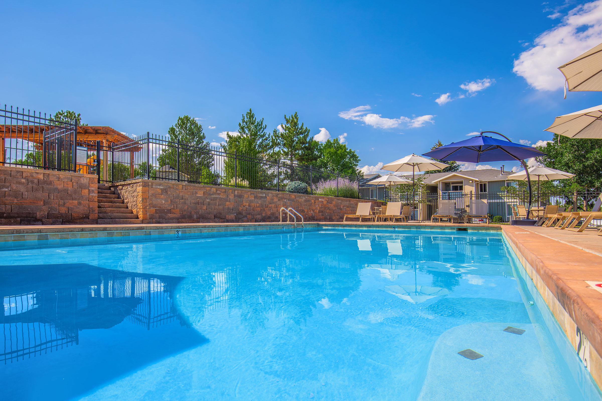 A clear blue swimming pool surrounded by lounge chairs and umbrellas, with a wooden pergola in the background. Tall trees and a bright blue sky with fluffy clouds add to the serene outdoor setting.