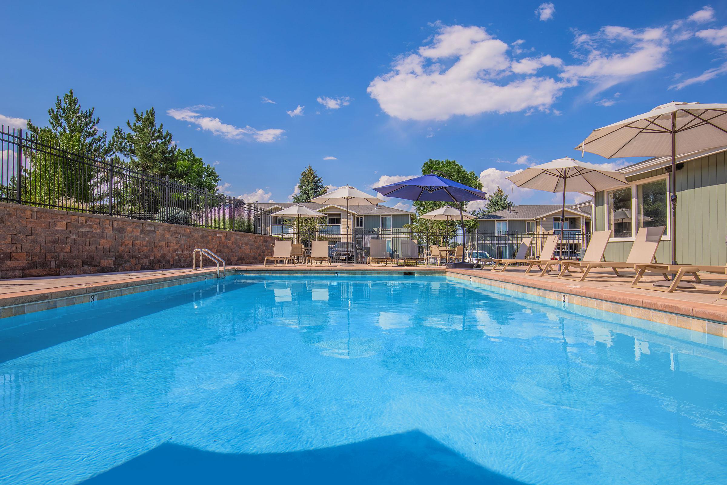 A clear blue swimming pool surrounded by lounge chairs and umbrellas on a sunny day. In the background, green trees and a fence can be seen under a bright blue sky with fluffy white clouds. The setting conveys a relaxing outdoor atmosphere.