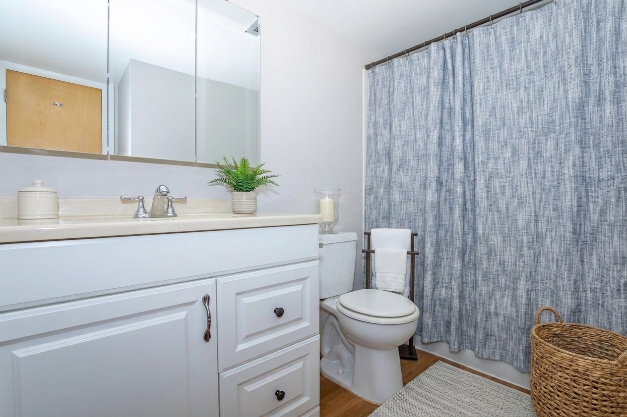 A clean, modern bathroom featuring a white vanity with a sink, a toilet, and a decorative shower curtain in a blue fabric. There is a wall mirror above the sink, a plant on the countertop, a rolled towel on the side, and a woven basket nearby. The floor is covered with a light-colored rug.
