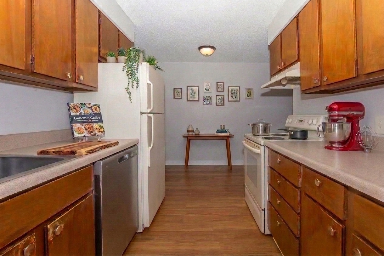 A modern kitchen featuring wooden cabinets, stainless steel appliances, and a countertop. There is a cookbook on the counter, a small table in the background with decorative frames, and a mixer on the counter beside the stove. The floor has a wood-like finish, creating a warm atmosphere.