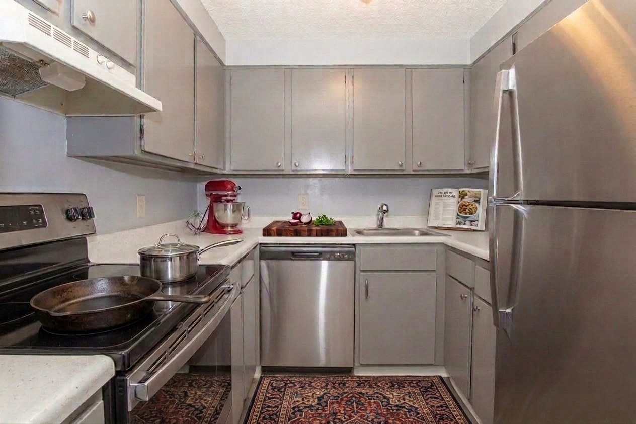 A modern kitchen featuring gray cabinets, stainless steel appliances, and a white countertop. An electric stove and oven are visible, along with a dishwasher and sink. A stand mixer and a small cutting board with vegetables are on the counter. A decorative rug is placed on the floor.