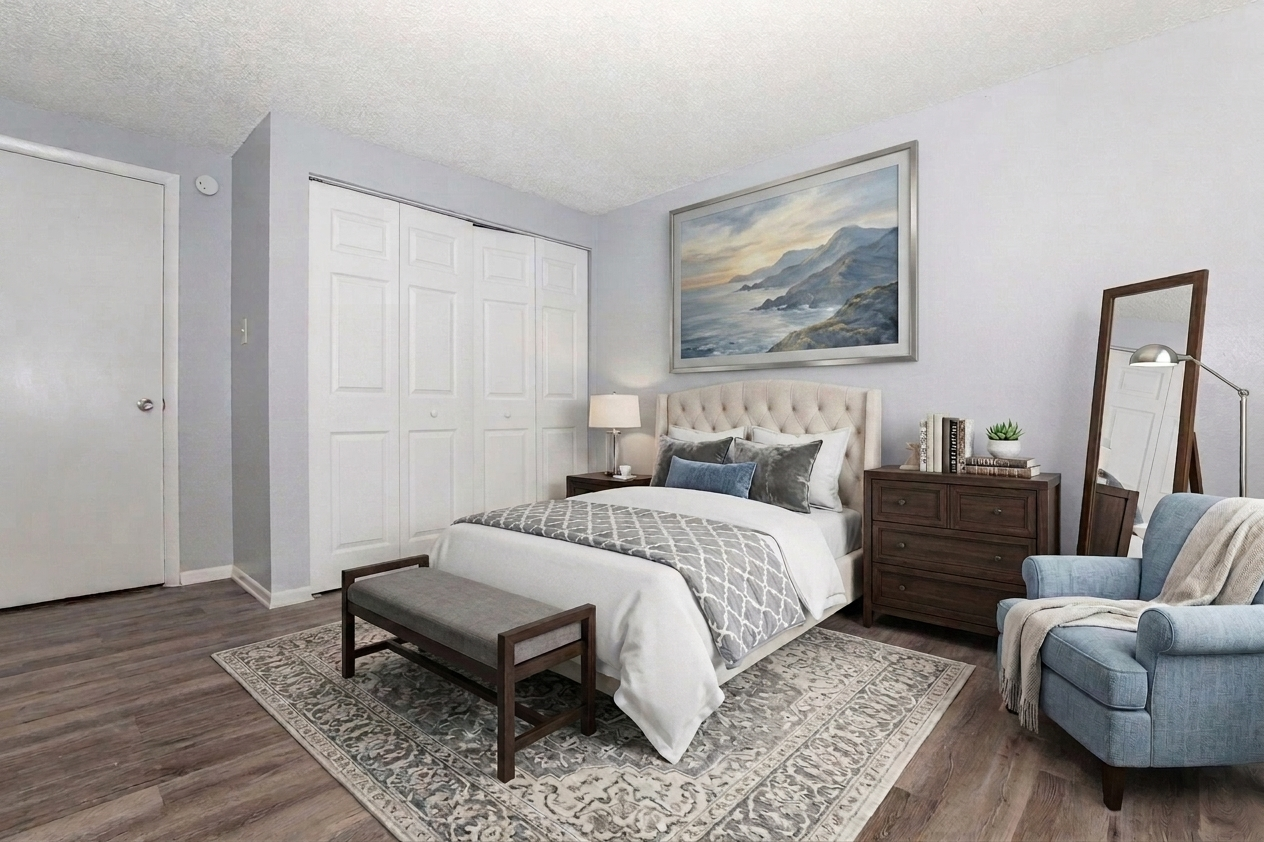 A cozy bedroom featuring a light gray color scheme. The room includes a bed with a tufted headboard, decorative pillows, a bedside lamp, and a wooden nightstand. A large painting of a landscape hangs on the wall, and there's a full-length mirror beside a blue armchair. The floor is wooden with a patterned area rug.