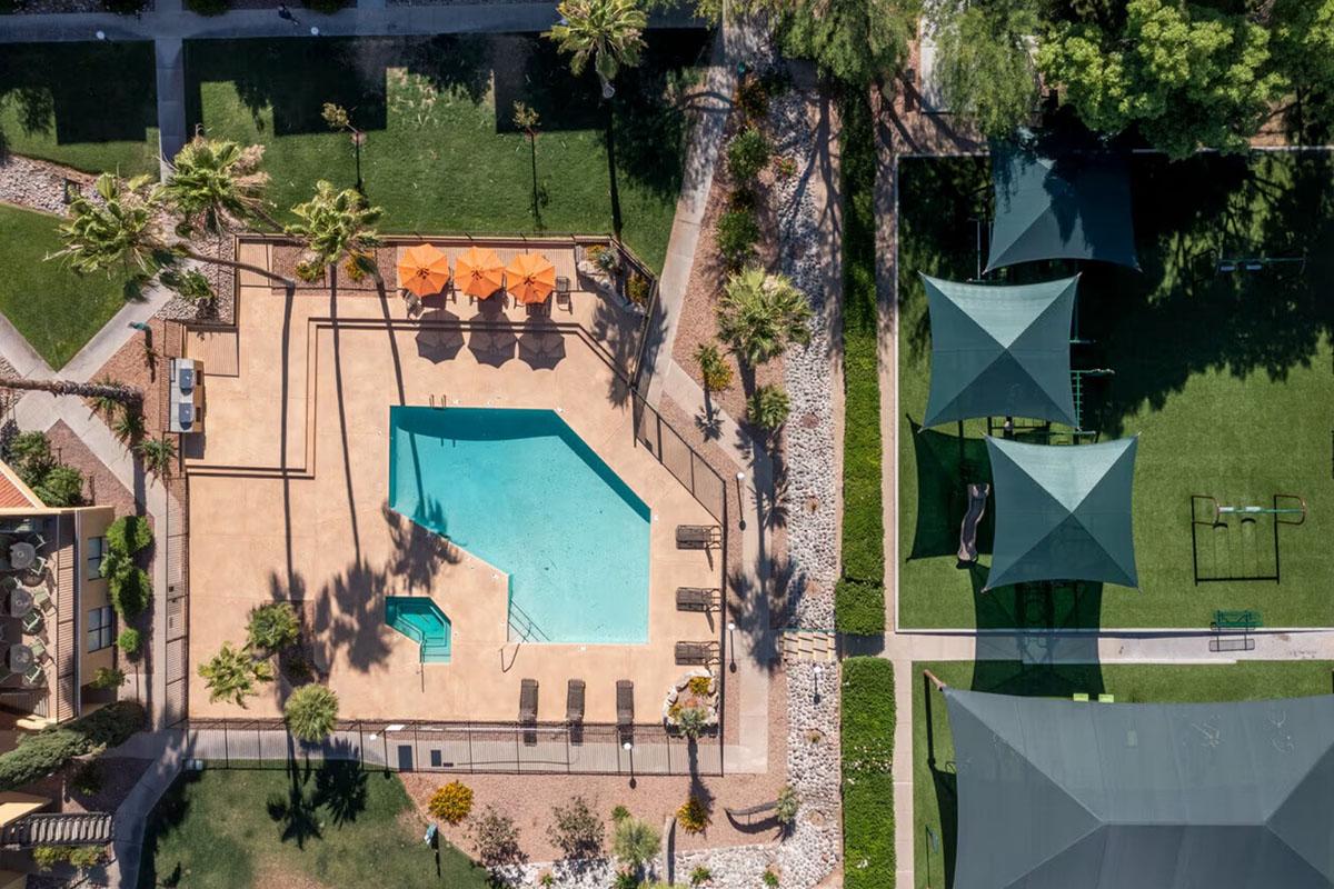 Aerial view of a swimming pool surrounded by lounge chairs and orange umbrellas, situated in a landscaped area with green grass and palm trees. Nearby, shaded structures and walking paths are visible, creating a tranquil outdoor environment.