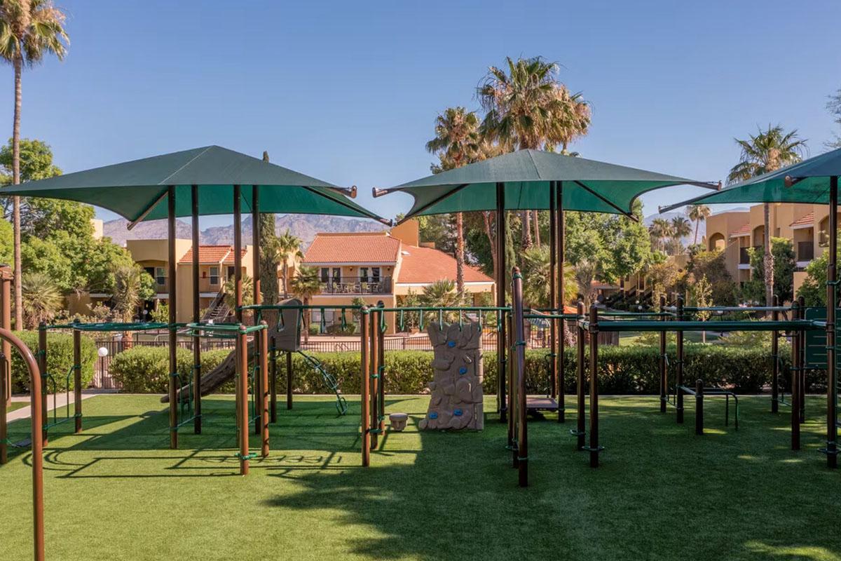 A playground featuring green canopies, climbing structures, and a rock wall, set in a sunny outdoor area with palm trees and residential buildings in the background.