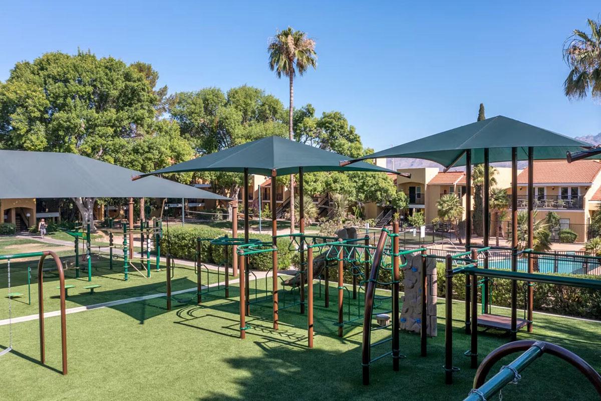 A playground area featuring green equipment and shaded structures. The playground is surrounded by trees and palm trees, with nearby buildings visible in the background. The grassy surface provides a safe play environment, creating a recreational space for children and families.