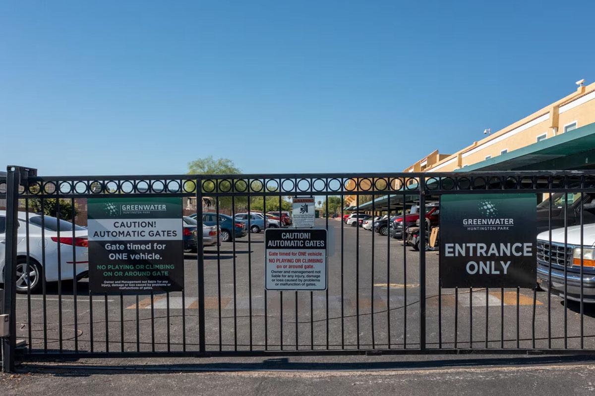 A gated entrance to a parking area, featuring signs that read "CAUTION! AUTOMATIC GATES" and "ENTRANCE ONLY." The scene includes several parked vehicles and a clear blue sky above.