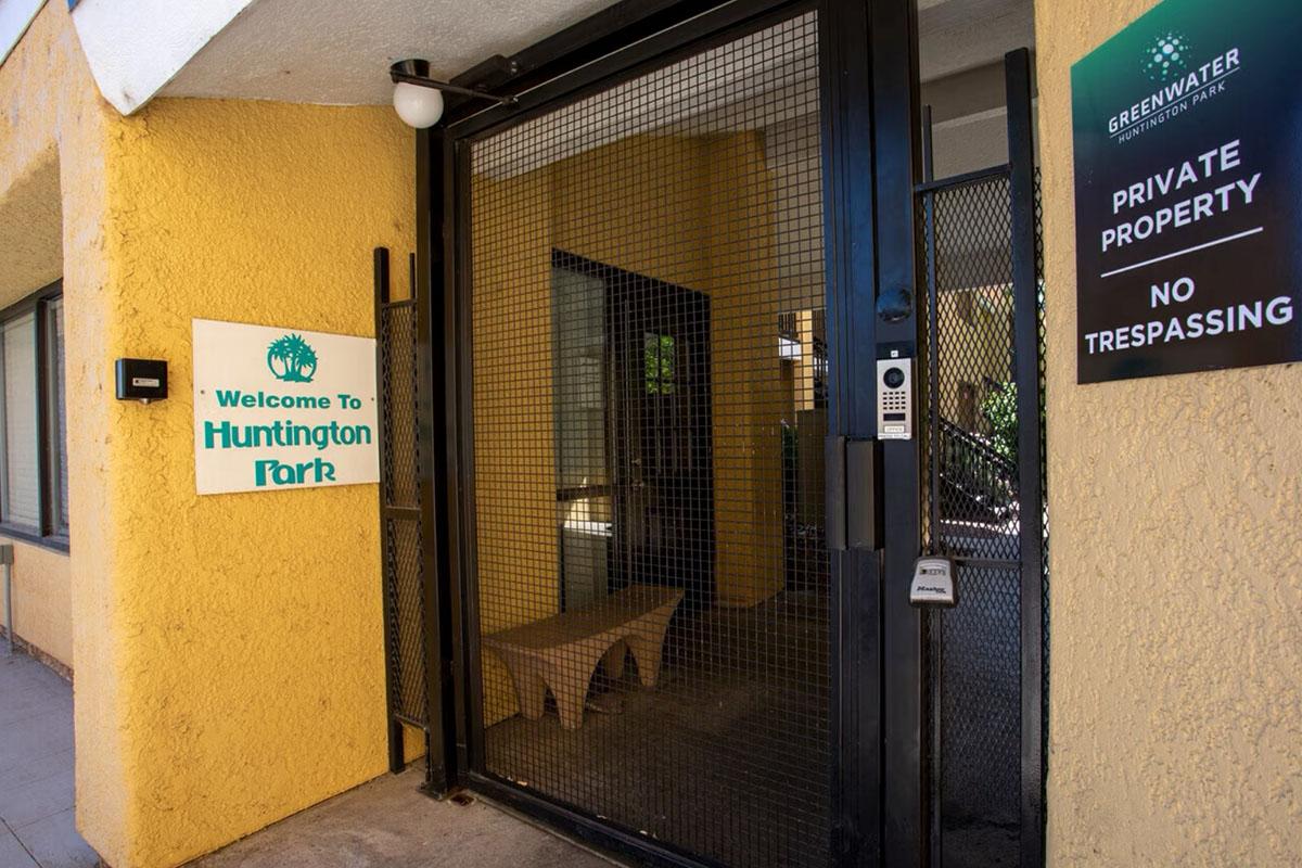 Entrance to Huntington Park with a sign stating "Welcome To Huntington Park." The gate is enclosed with a security mesh, and there are notices indicating "Private Property" and "No Trespassing." The building exterior is painted a light yellow, adding a welcoming yet secure appearance.
